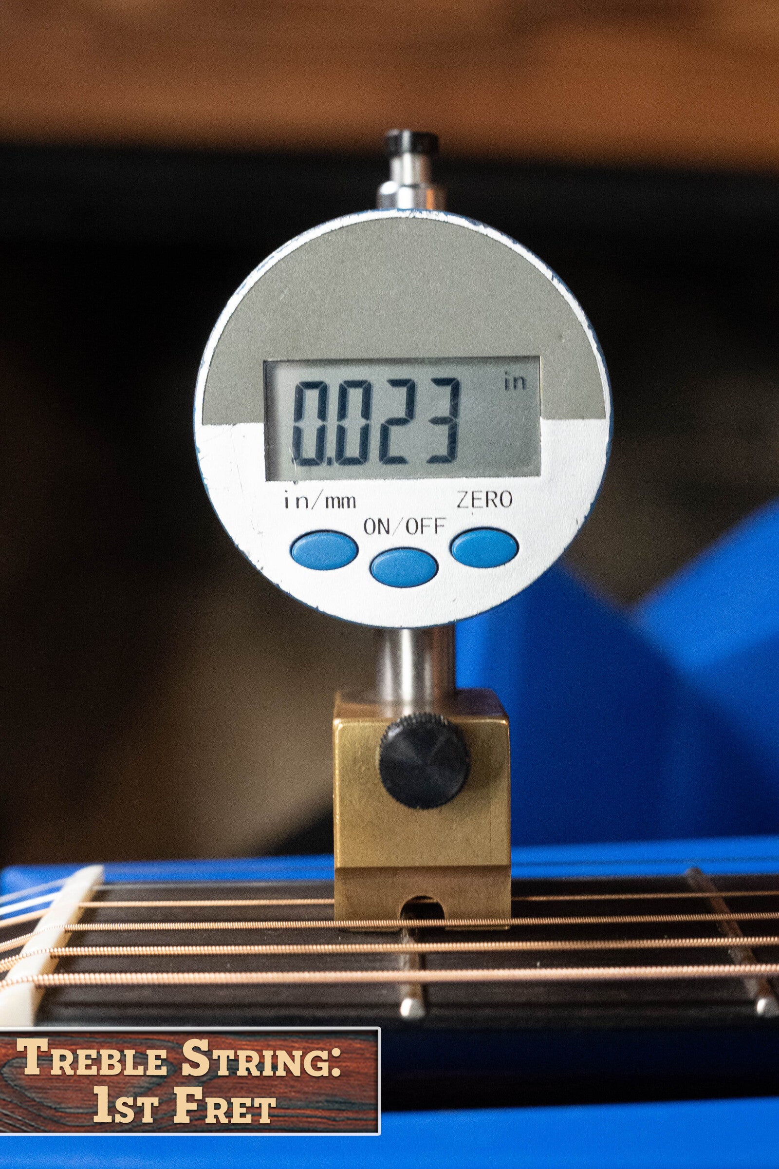 A digital caliper measures 0.023 inches above the first fret of a blue and gold Bourgeois Touchstone Series Dreadnought Acoustic Guitar #7014, with both resting near a label: Treble String: 1st Fret.