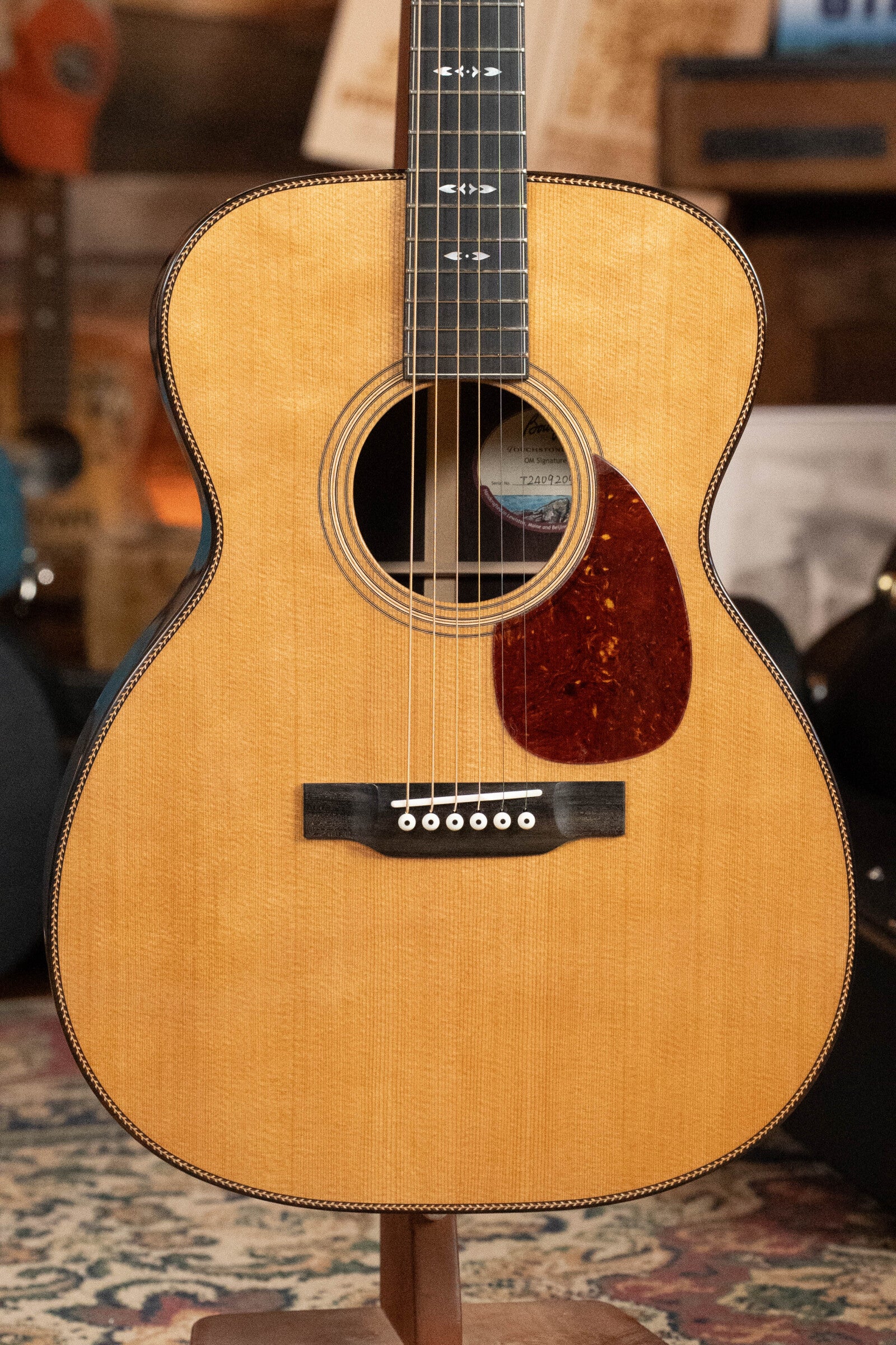 The Bourgeois OM Touchstone Signature Torrefied Alaskan Sitka/Madagascar Rosewood Acoustic Guitar #9209 stands on a guitar stand, showcasing its natural finish, dark inlay, tortoiseshell pickguard, and white bridge pins against a blurred backdrop.