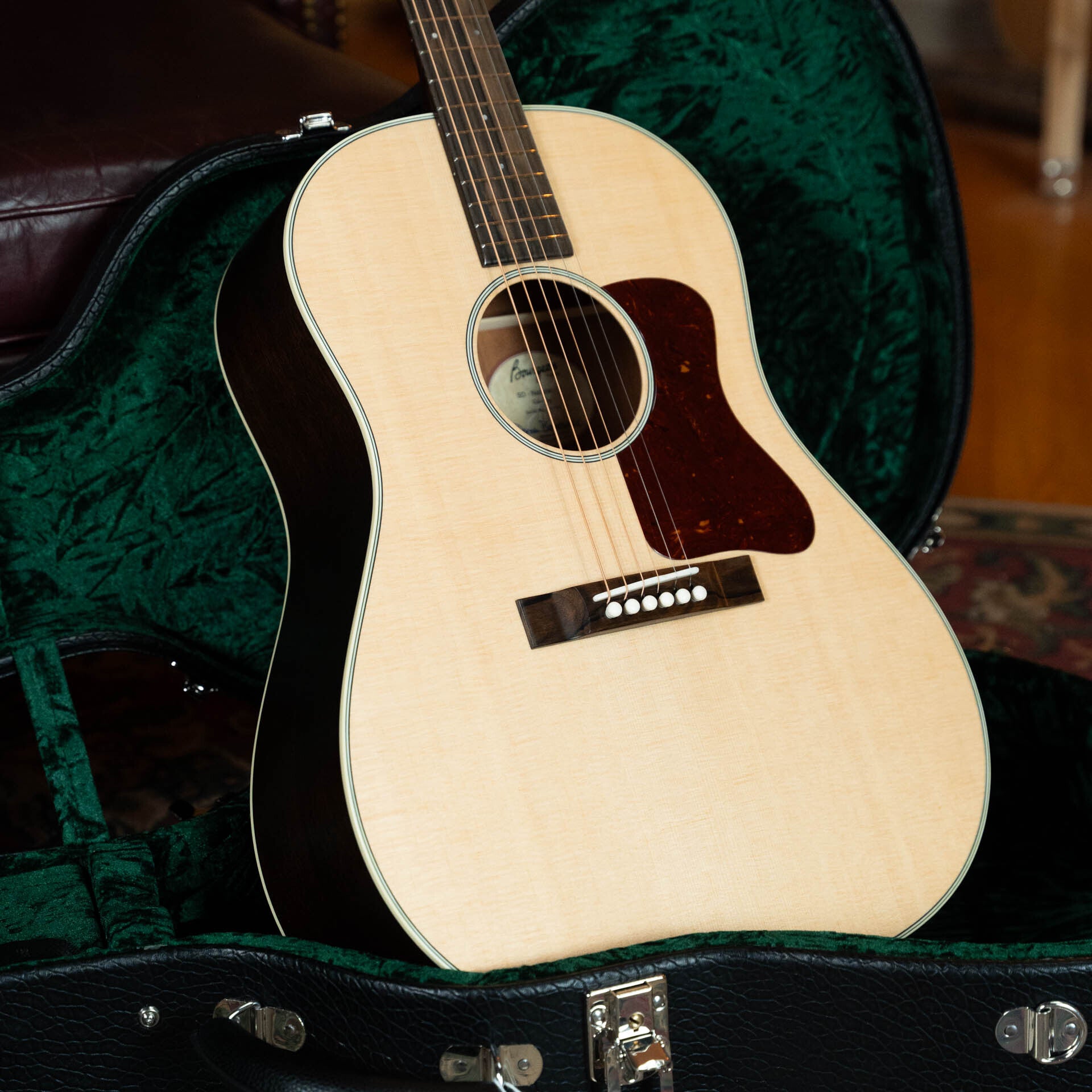A Boucher Guitars PSN-SD-S Satin Professional Series The Natural Sitka/Mahogany Slope Shoulder Dreadnought Acoustic Guitar #1174 with a dark pickguard is shown in an open, green-lined hard case, with furniture and carpet in the background.