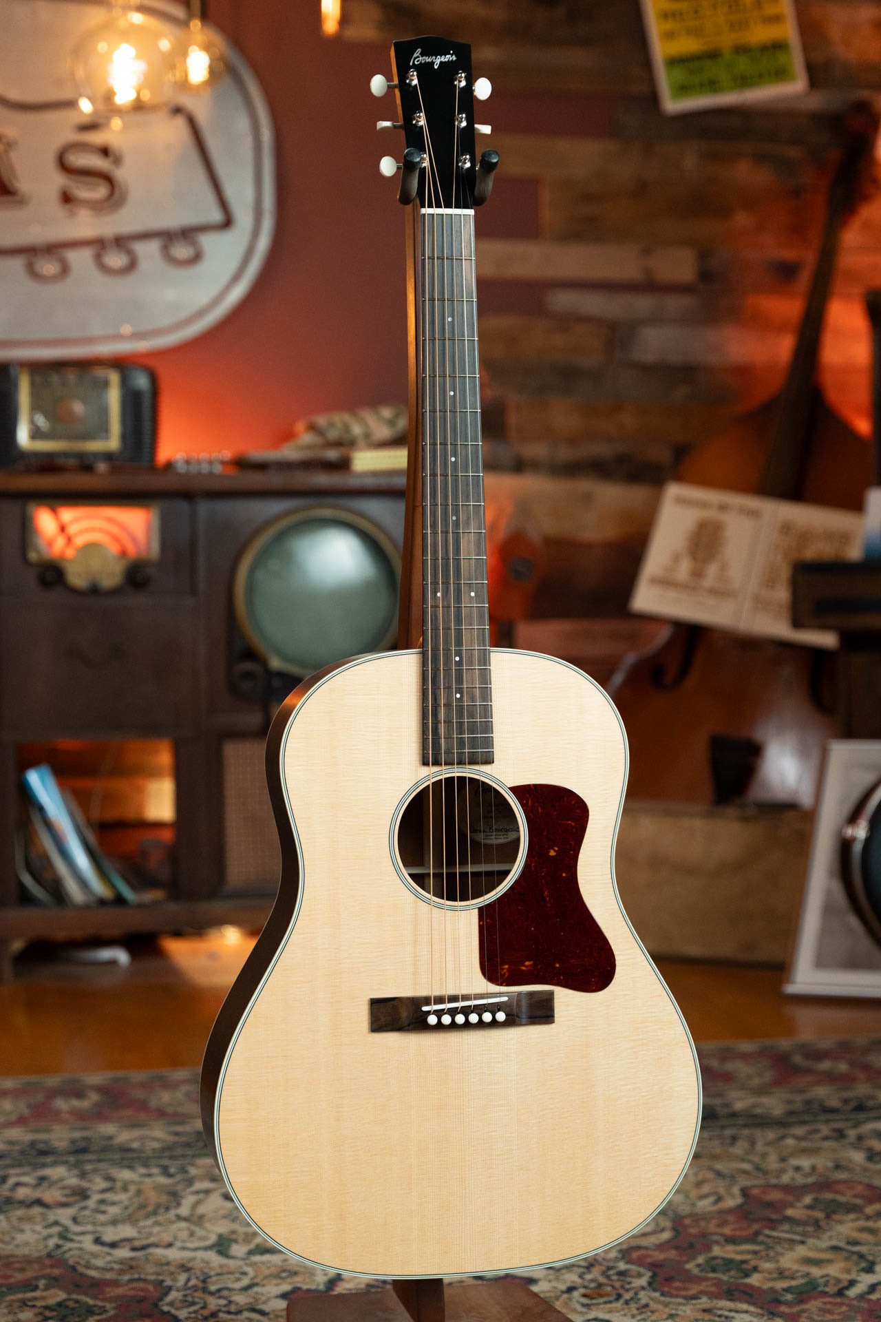 A Boucher Guitars PSN-SD-S Satin Professional Series Sitka/Mahogany Slope Shoulder Dreadnought Acoustic Guitar stands upright on a patterned rug in a cozy, vintage-style room with wood-paneled walls and retro decor.