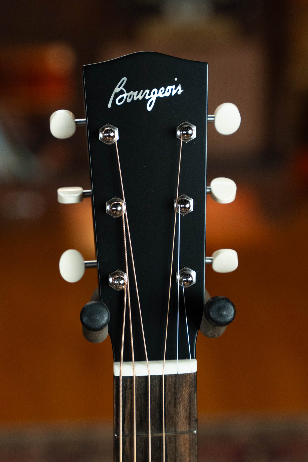 Close-up of the Boucher Guitars Bourgeois PSN-SD-S Satin Professional Series Slope Shoulder Dreadnought Acoustic Guitar #1174 headstock in black with white script, six tuners, and two supports, set against a blurred brown background.