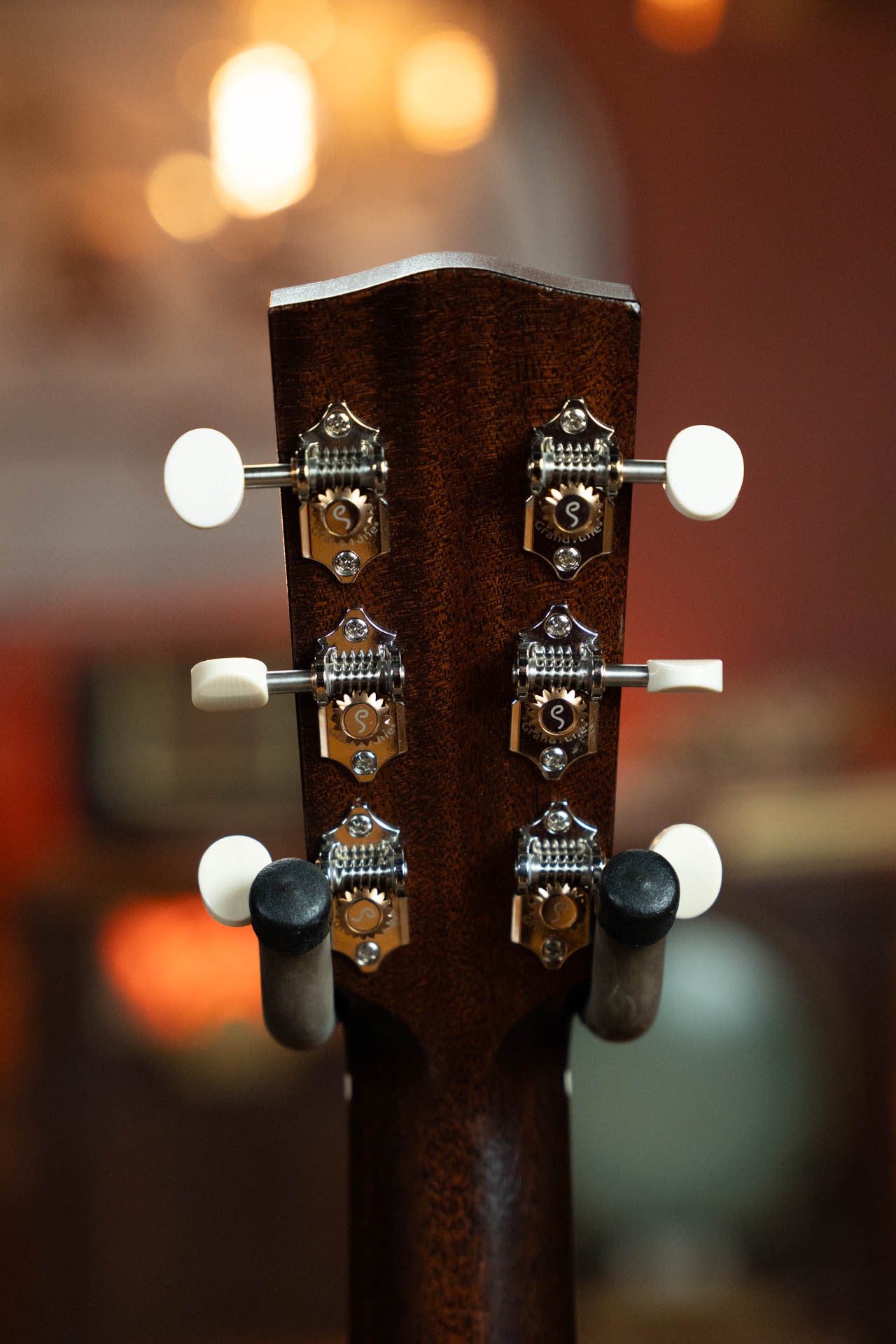 Close-up of the back of a Boucher Guitars Bourgeois PSN-SD-S Satin Professional Series Slope Shoulder Dreadnought Acoustic Guitar #1174 headstock, showing six metal tuning pegs with white knobs against a warmly lit, blurred background.
