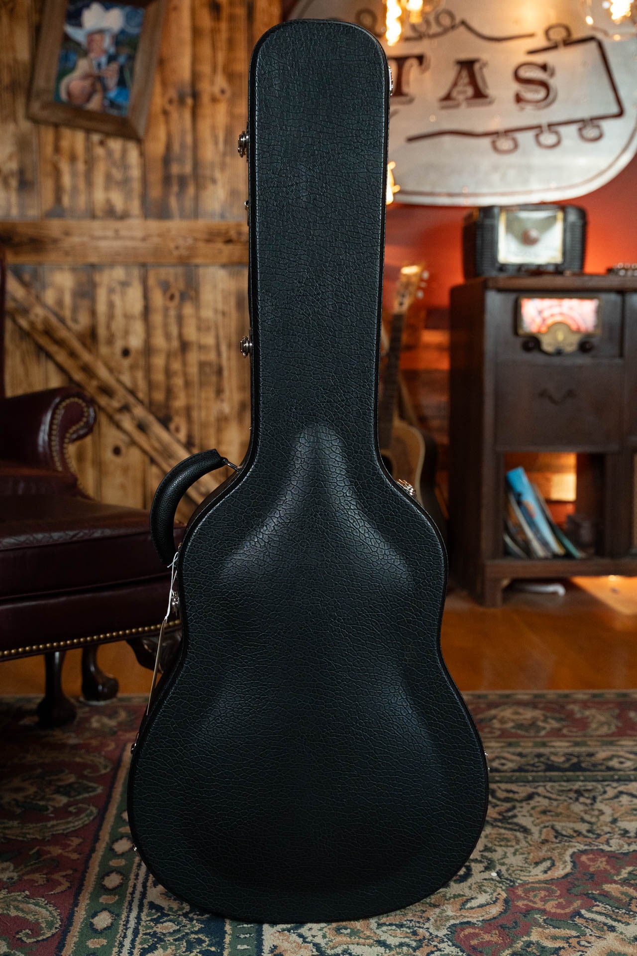 A black guitar case, likely holding a Boucher Guitars Bourgeois PSN-SD-S Satin Professional Series The Natural Sitka/Mahogany Slope Shoulder Dreadnought Acoustic Guitar #1174, stands upright on a patterned rug in a cozy room with wooden walls and warm lighting.