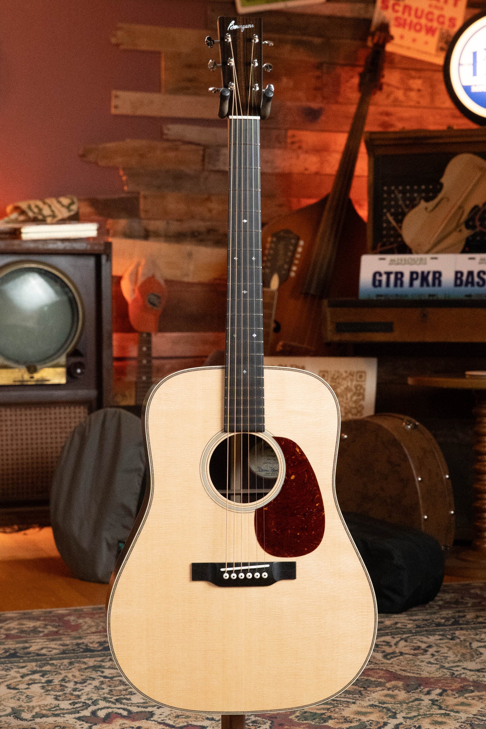 A Bourgeois PSV-D-S Professional Series Sitka/Rosewood Vintage Satin Dreadnought Acoustic Guitar #1124 stands upright in a cozy room with vintage musical equipment and wooden walls.