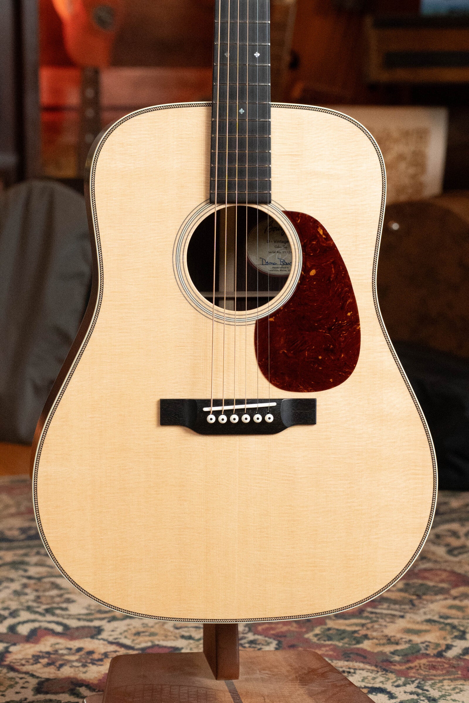 A close-up of the Bourgeois PSV-D-S Professional Series Sitka/Rosewood Vintage Satin Dreadnought Acoustic Guitar #1124 stands upright on a wooden stand atop a patterned rug, showcasing its six strings and dark pickguard.
