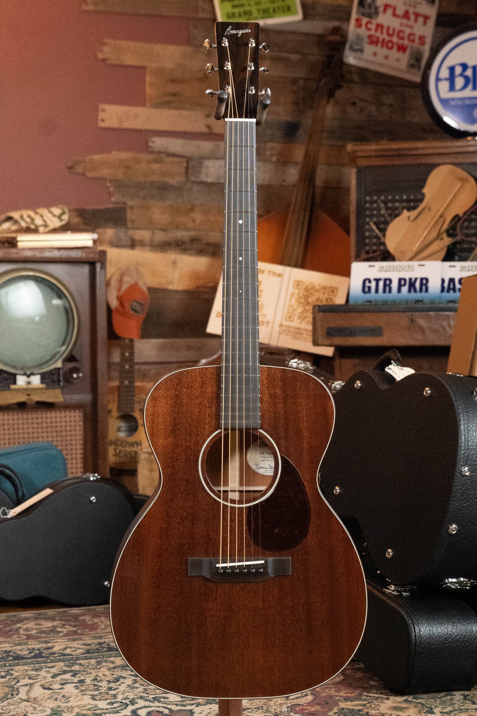 A Bourgeois Professional Series Hog Top All Mahogany OM Acoustic Guitar #1208 by Bourgeois stands upright in a cozy, vintage-style room with a TV, guitar cases, and retro decor in the background.