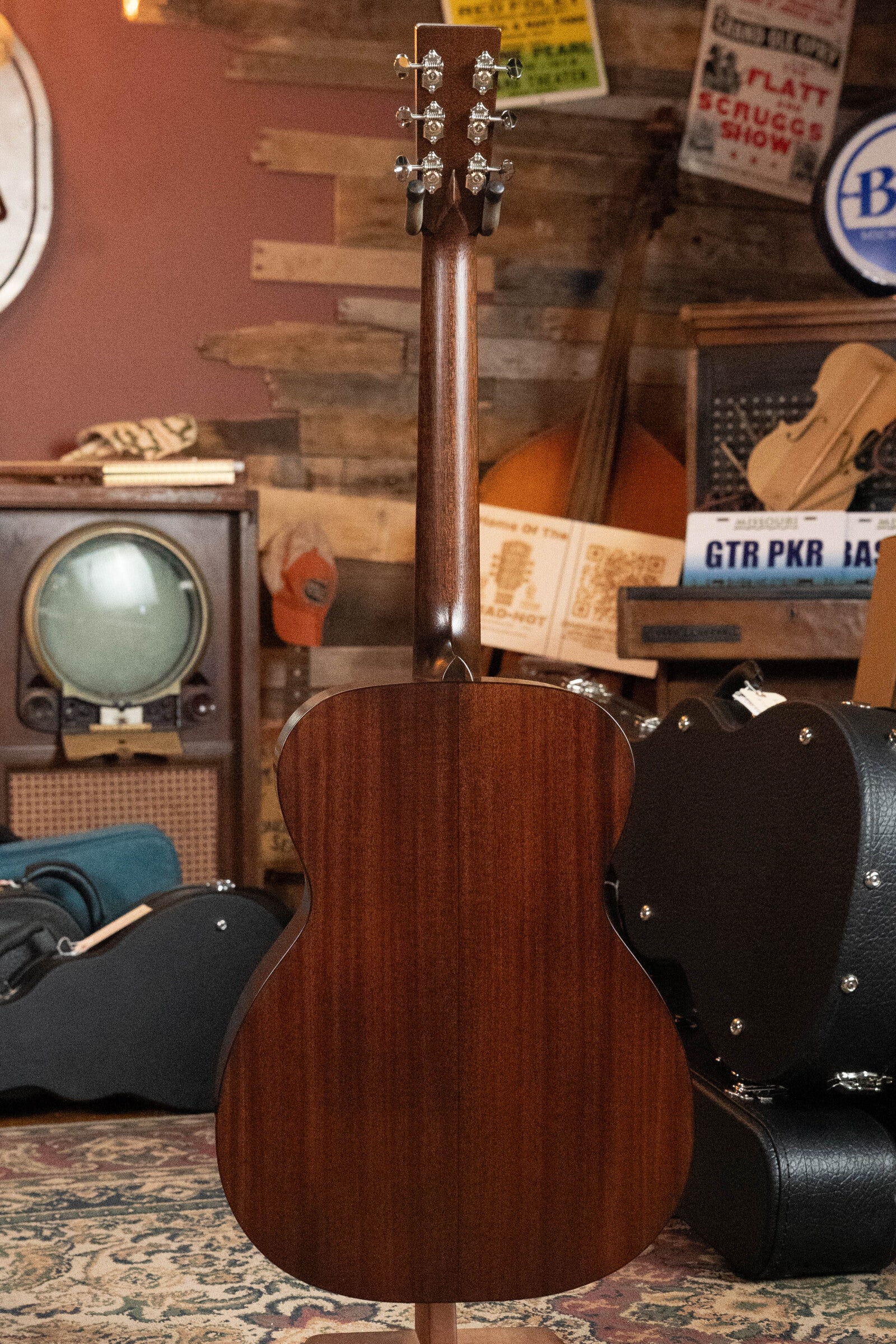 The Bourgeois Professional Series Hog Top All Mahogany OM Acoustic Guitar #1208 by Bourgeois is pictured from the back, standing upright in a vintage-themed room with a retro TV, instrument cases, and wooden wall panels.