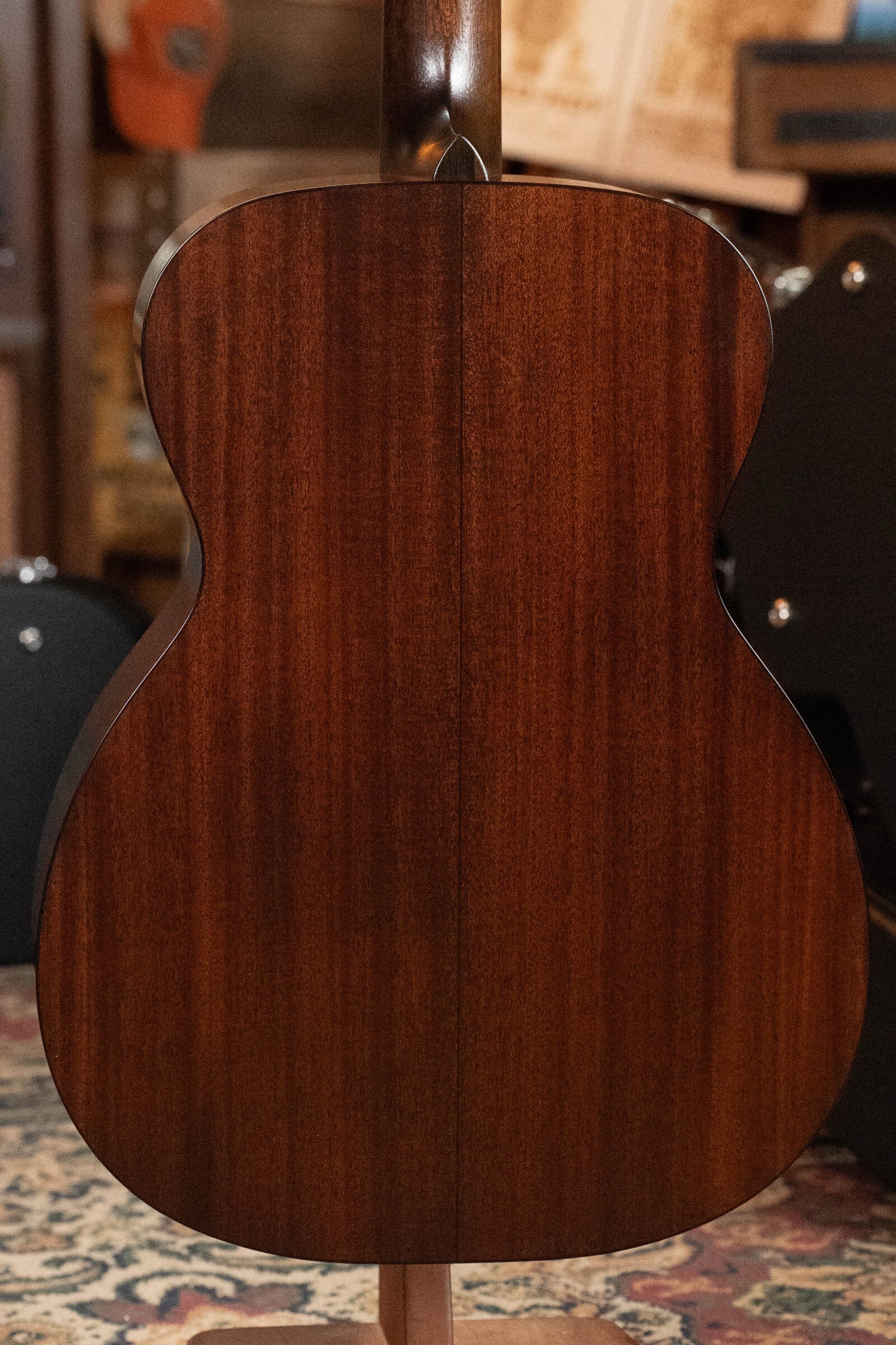 The image shows the back of a Bourgeois Professional Series Hog Top All Mahogany OM Acoustic Guitar #1208 standing upright indoors, with blurred musical instruments and cases visible in the background.