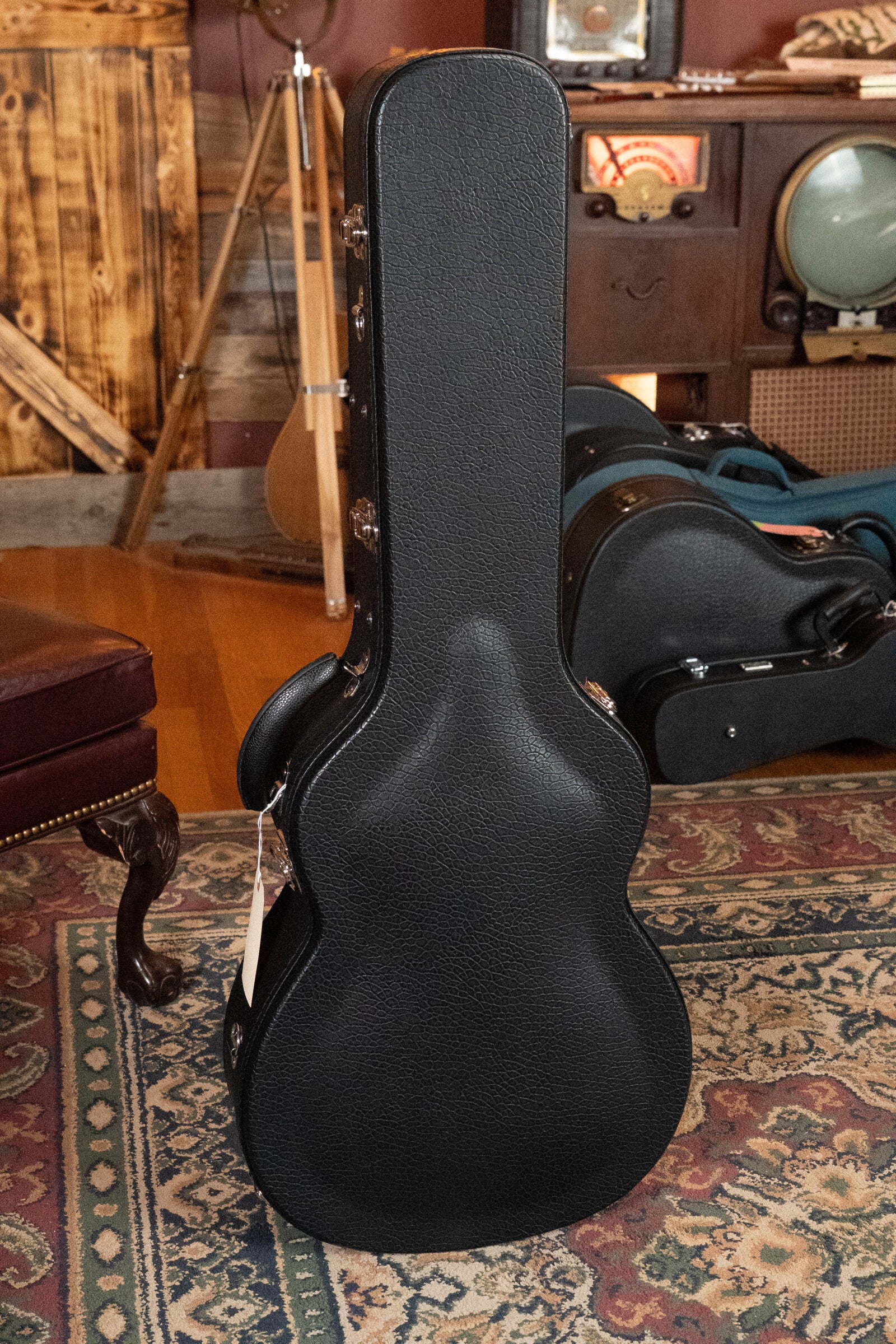 A black hard-shell case stands upright on a patterned rug in a cozy, vintage-inspired room—possibly protecting the Bourgeois Professional Series Hog Top All Mahogany OM Acoustic Guitar #1208.