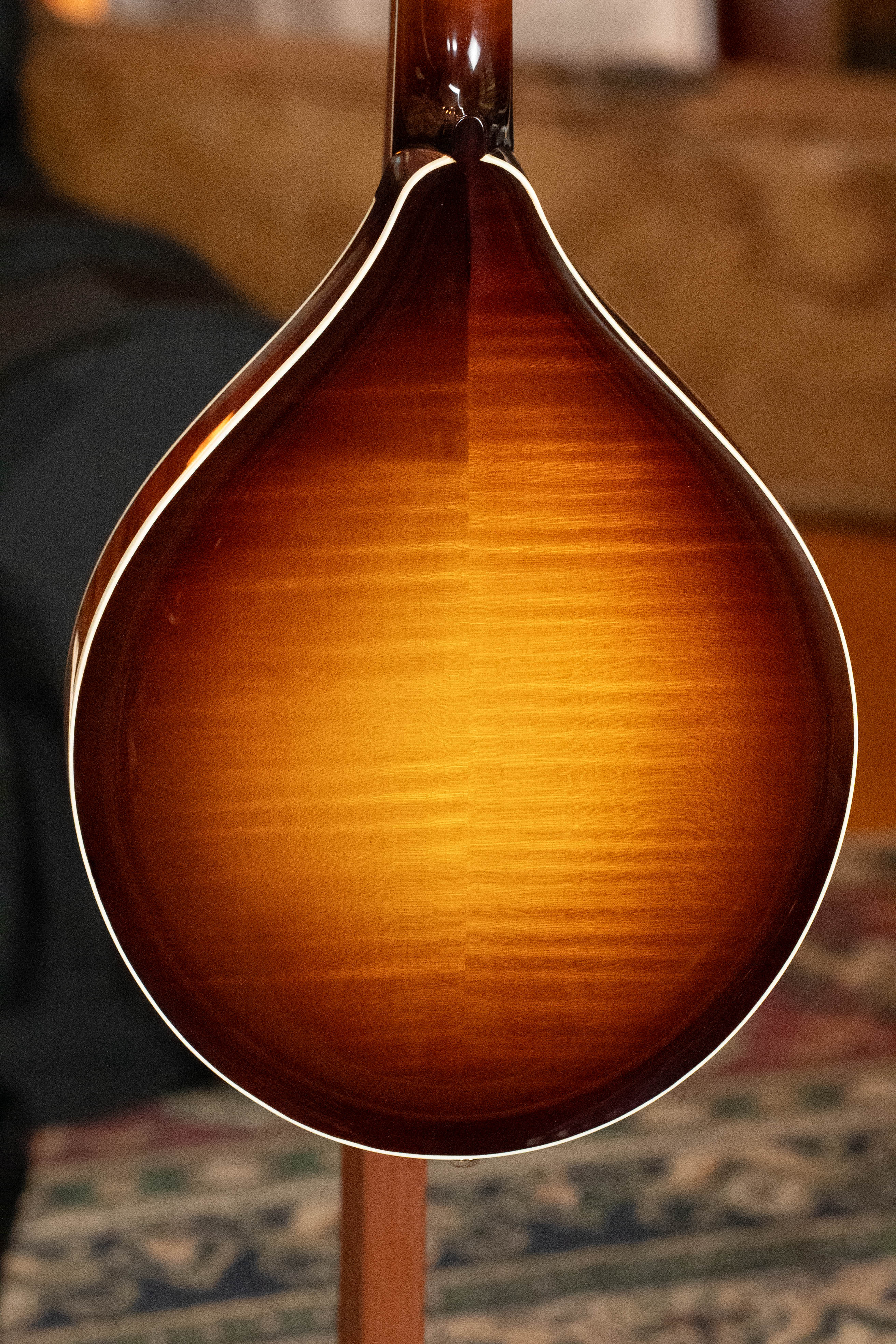 A close-up of the Bulas Carpathian Spruce/Curly Maple Cremona Sunburst A-Style Mandolin #8129 shows its glossy teardrop back and light wood grain, set against a blurred indoor background.