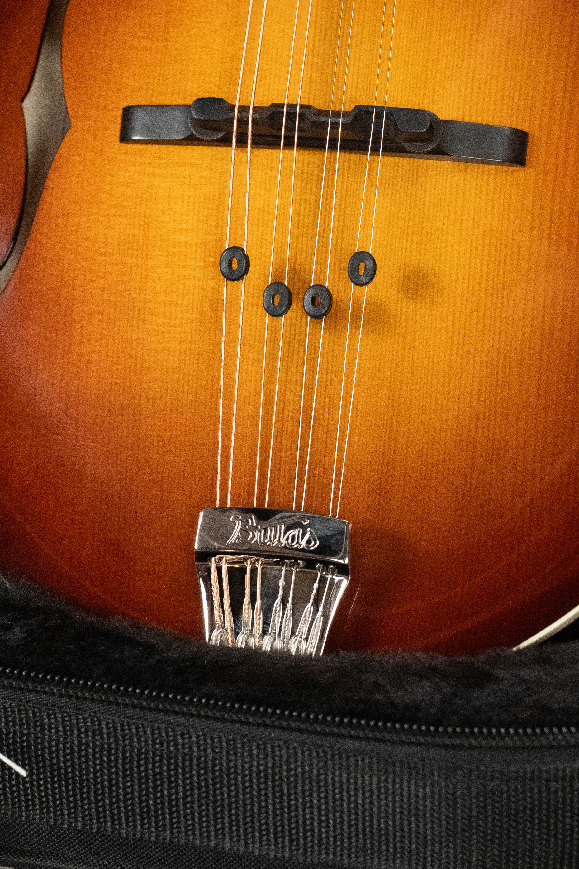 Close-up of the Bulas Carpathian Spruce/Curly Maple Cremona Sunburst A-Style Mandolin #8129, showing its strings, bridge, and branded chrome tailpiece inside a black fur-lined case.