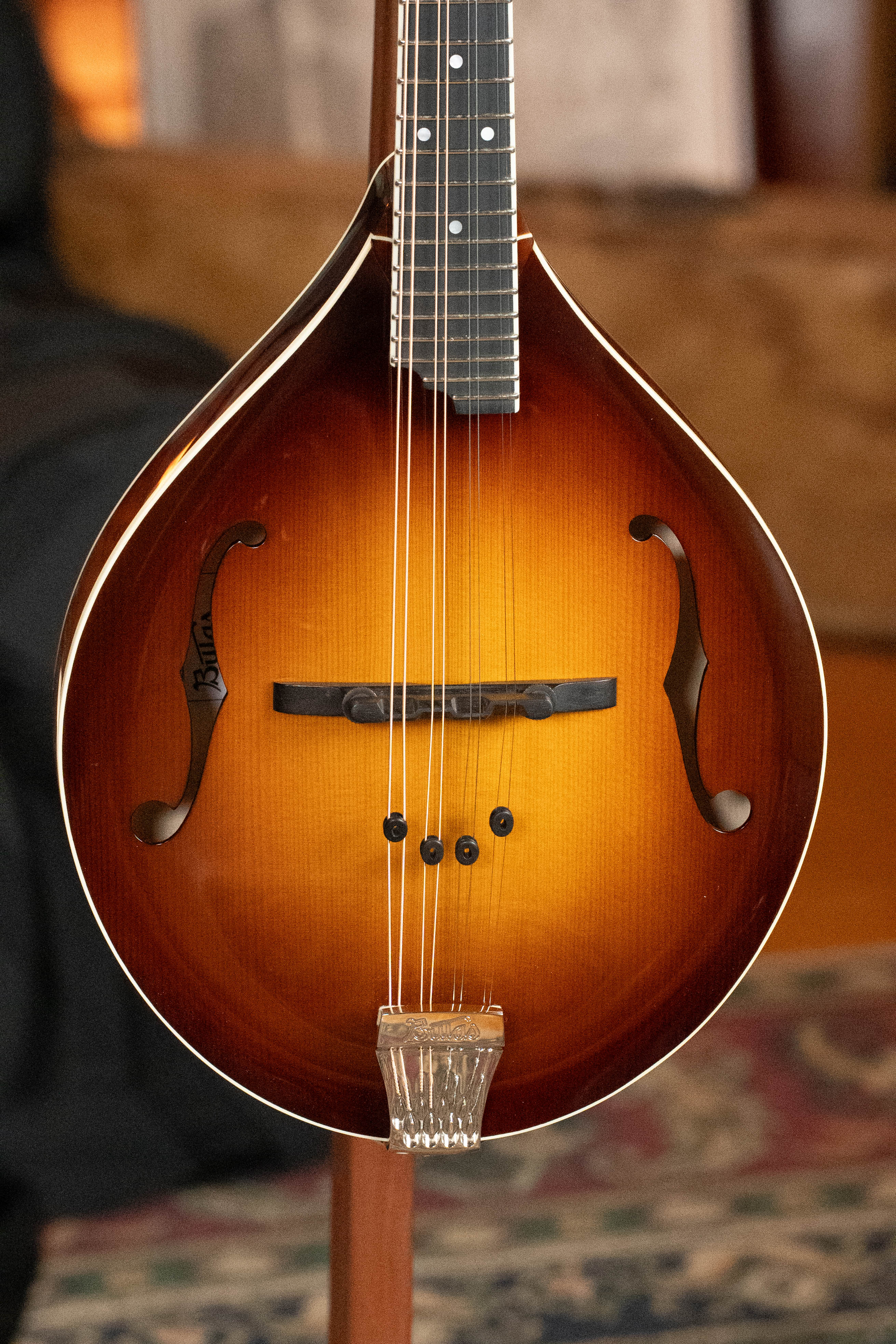 The Bulas Carpathian Spruce/Curly Maple Cremona Sunburst A-Style Mandolin #8129 stands upright on a patterned rug, showing off its f-holes and wooden bridge, with furniture and books softly blurred in the background.