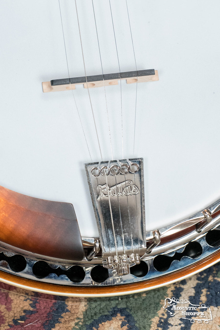 Close-up of the Bulas 5 String Resonator Banjo #0767 by Bulas, showing its strings, bridge, engraved metal tailpiece, part of the maple rim, and a decorative rug in the background.