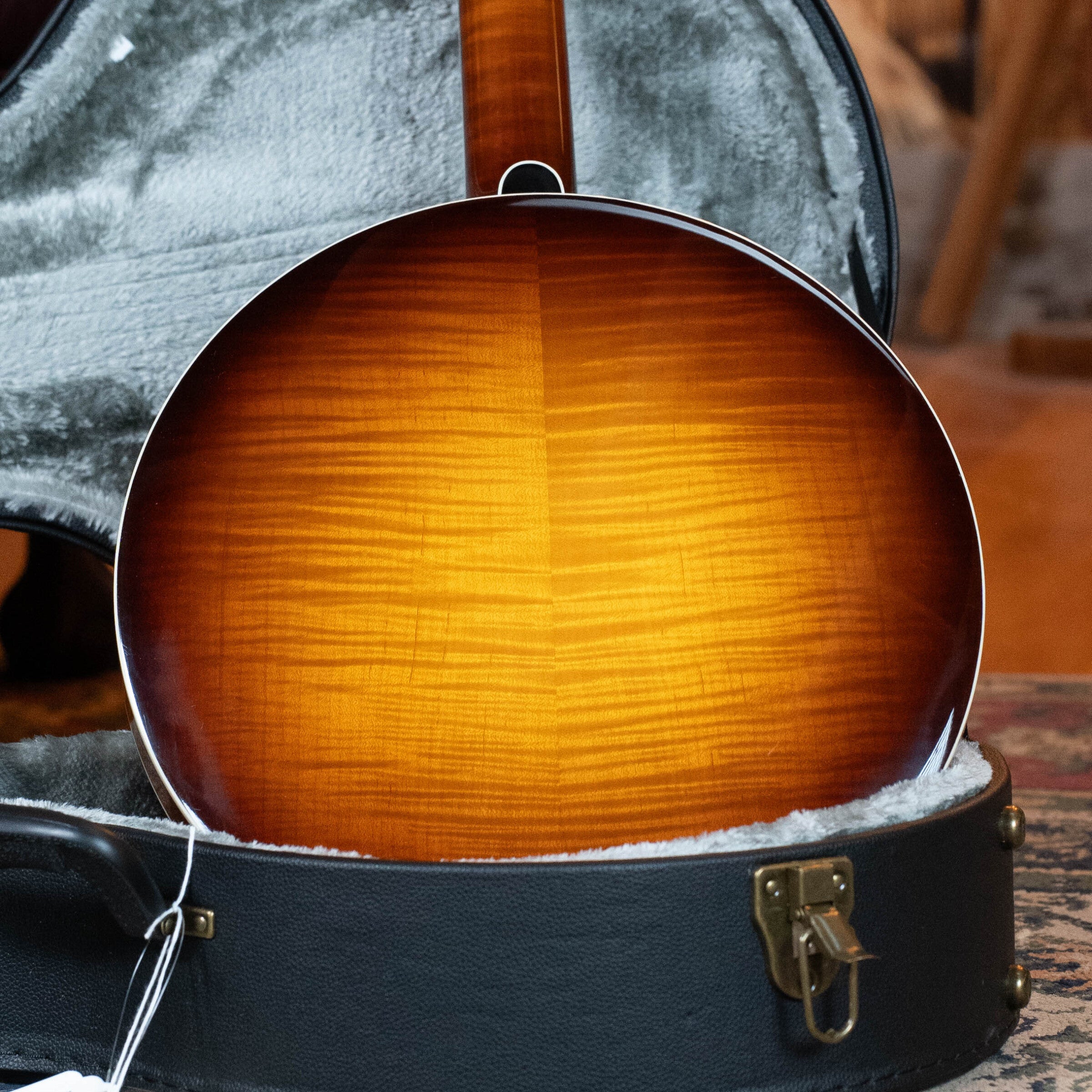 A close-up of the Bulas Standard 5-String Tobacco Sunburst Resonator Banjo #0676 shows its glossy, wood-grained back in a grey-lined open hard case with a textured interior and brass latch, highlighting Bulas craftsmanship.