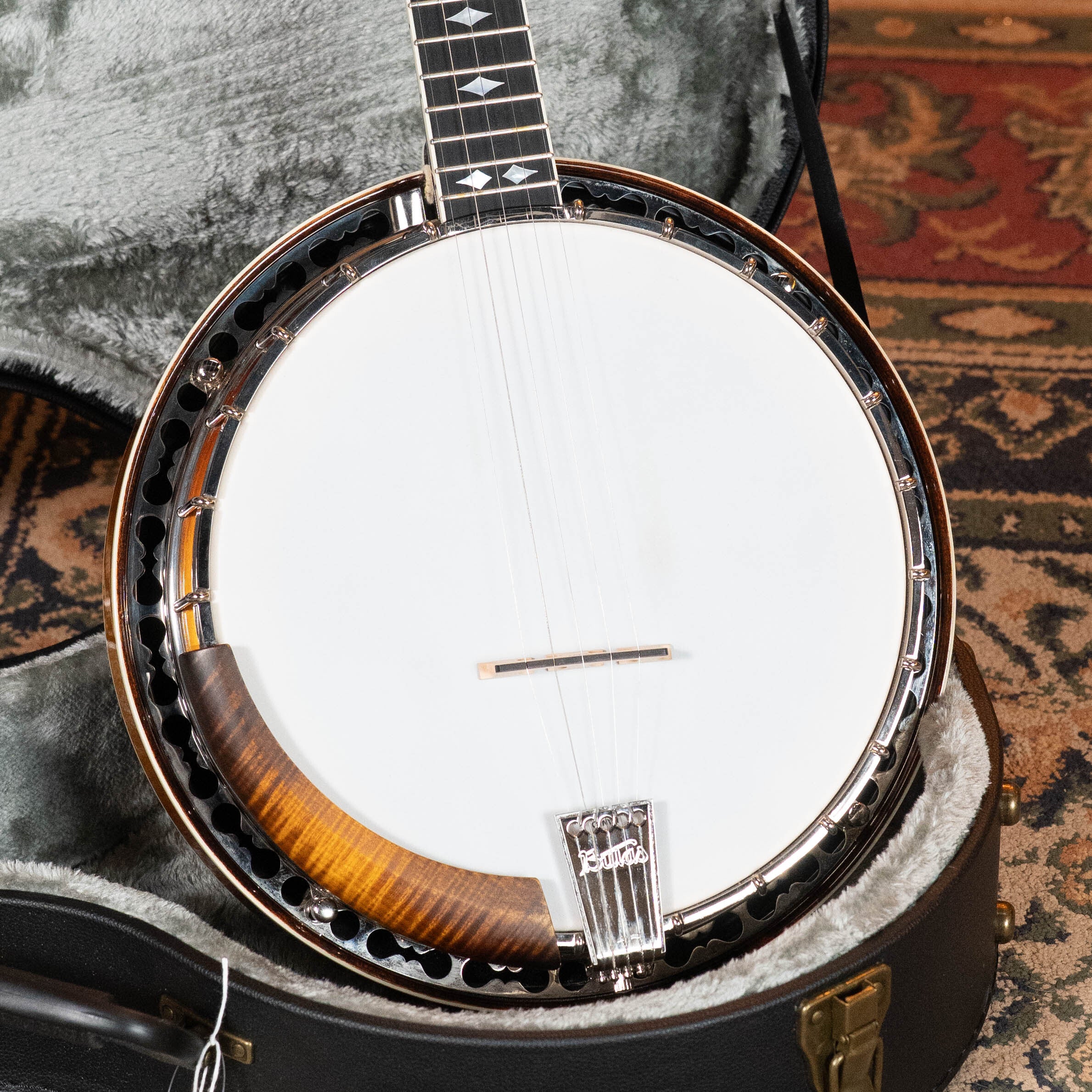A close-up of the Bulas Standard 5-String Tobacco Sunburst Resonator Banjo #0676 by Bulas, resting in an open padded case on a patterned rug, highlights its white drumhead, wooden rim, and metal hardware.