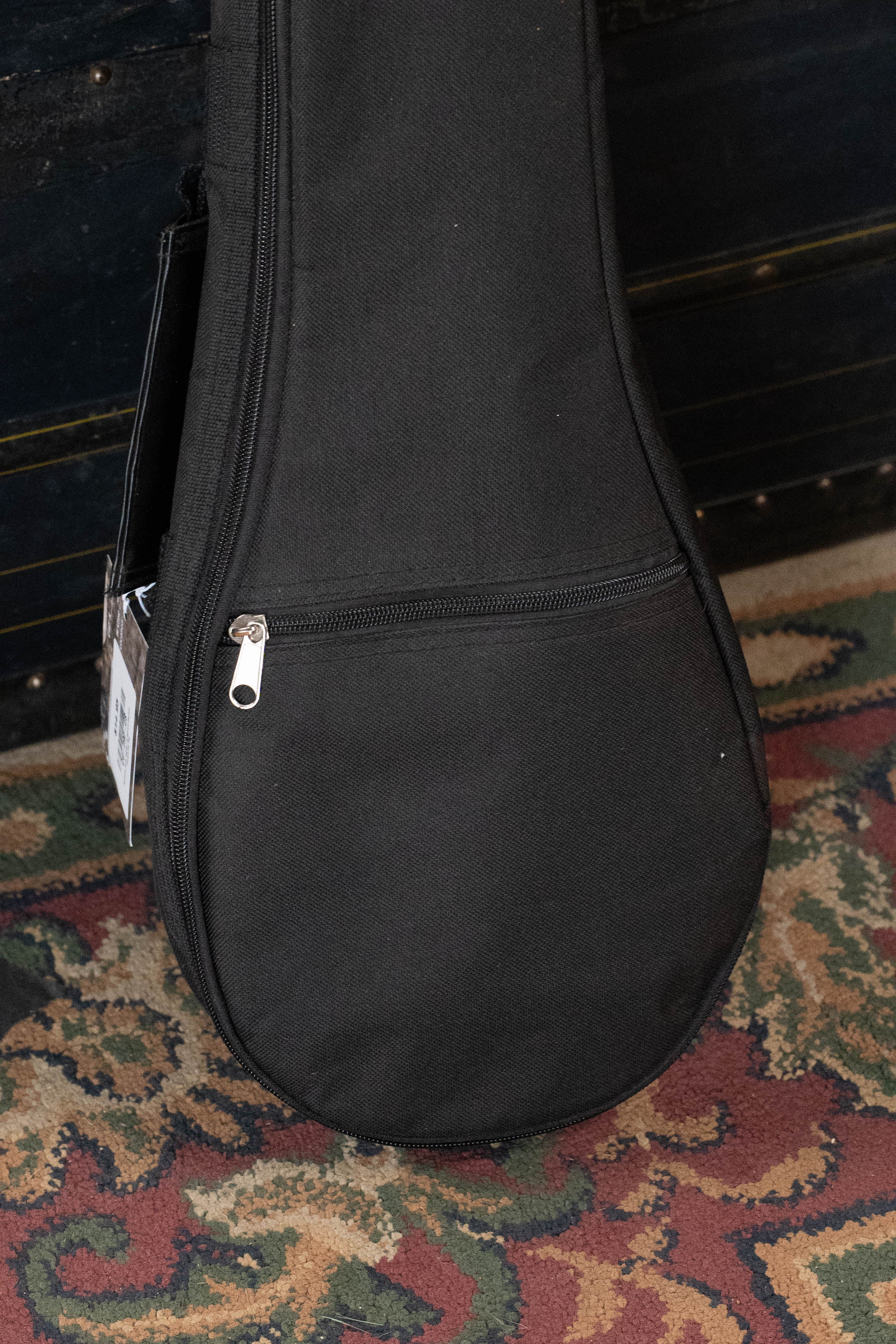 The Guardian 6mm Padded Duraguard Soprano Ukulele Gig Bag by Guardian, in black fabric with a zippered pocket, sits on a patterned carpet with part of a dark wooden surface seen in the background.