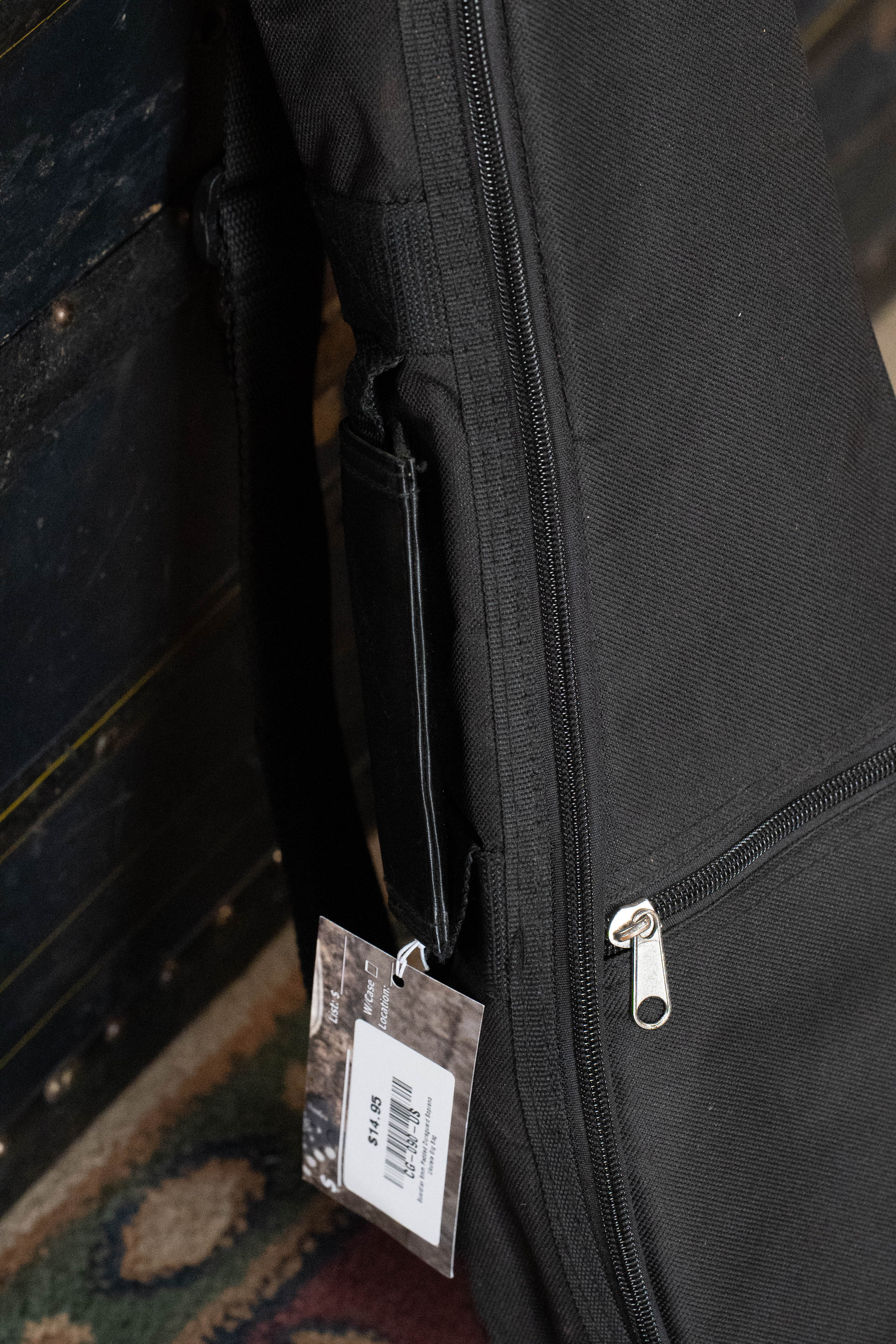 A close-up of the Guardian 6mm Padded Duraguard Soprano Ukulele Gig Bag, black with high-density padding and a zipper, featuring a white price tag, resting on a patterned carpet near a dark wooden chest.