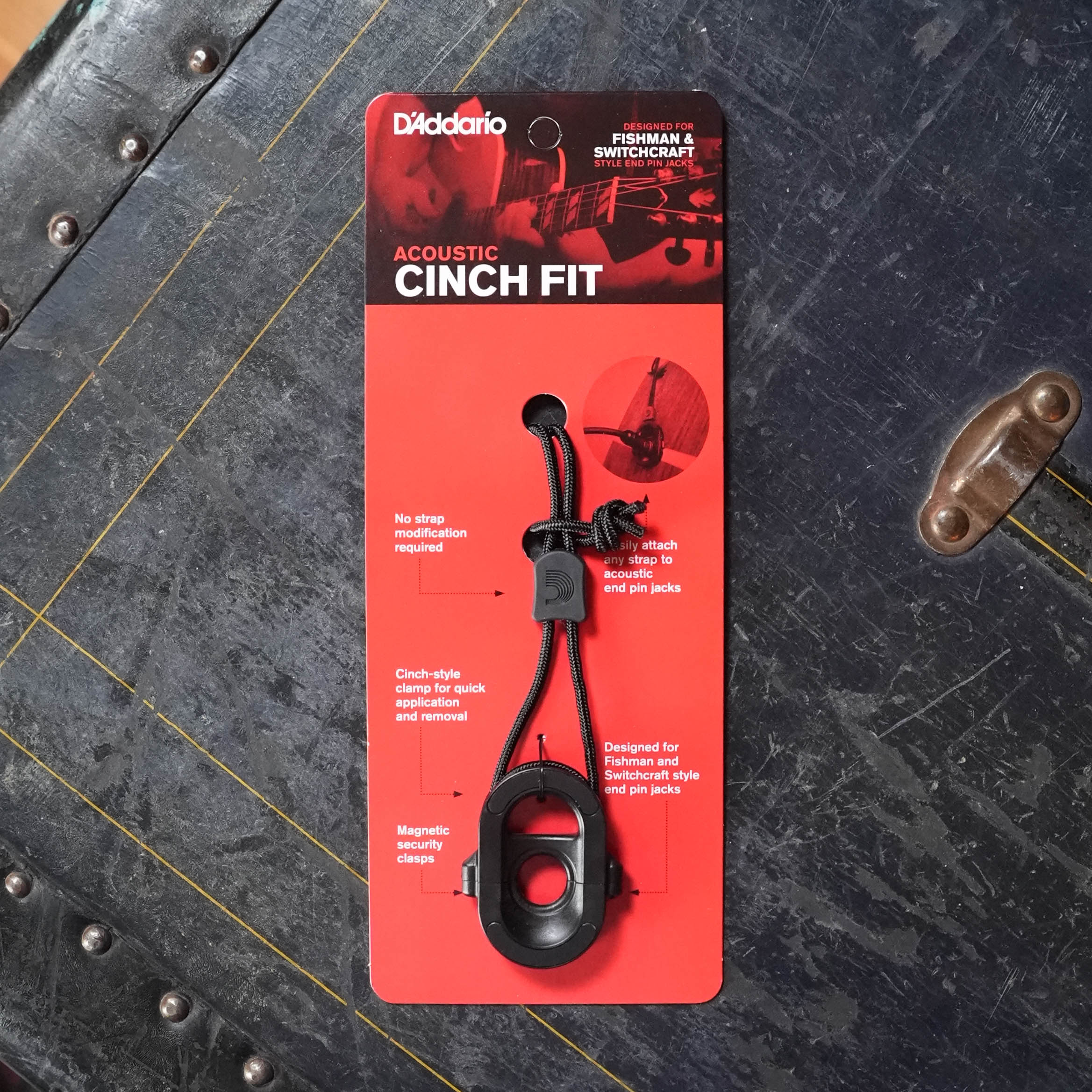 A D'Addario PW-AJL-01 CinchFit: Acoustic Jack Lock, a black guitar security device, is in its red and black packaging on a dark textured surface. The packaging displays key features and product details.