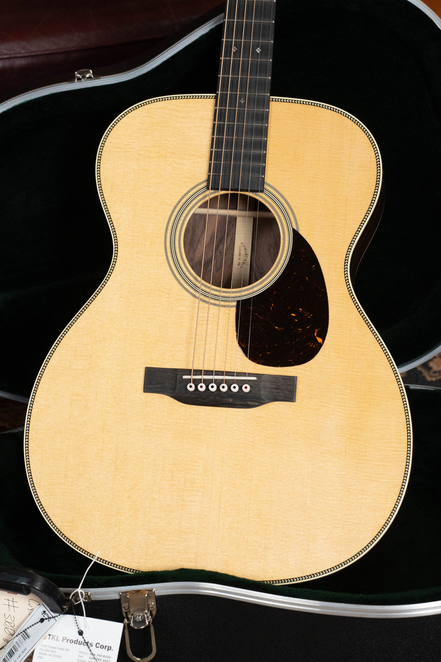 A close-up of the Martin & Co. Custom Shop OOO-28 Style "Wild Grain" East Indian Rosewood Spruce/Indian Rosewood Custom "OOO/OM" Acoustic Guitar, showing its light wood finish in a black velvet-lined hard case with tags near the bottom.