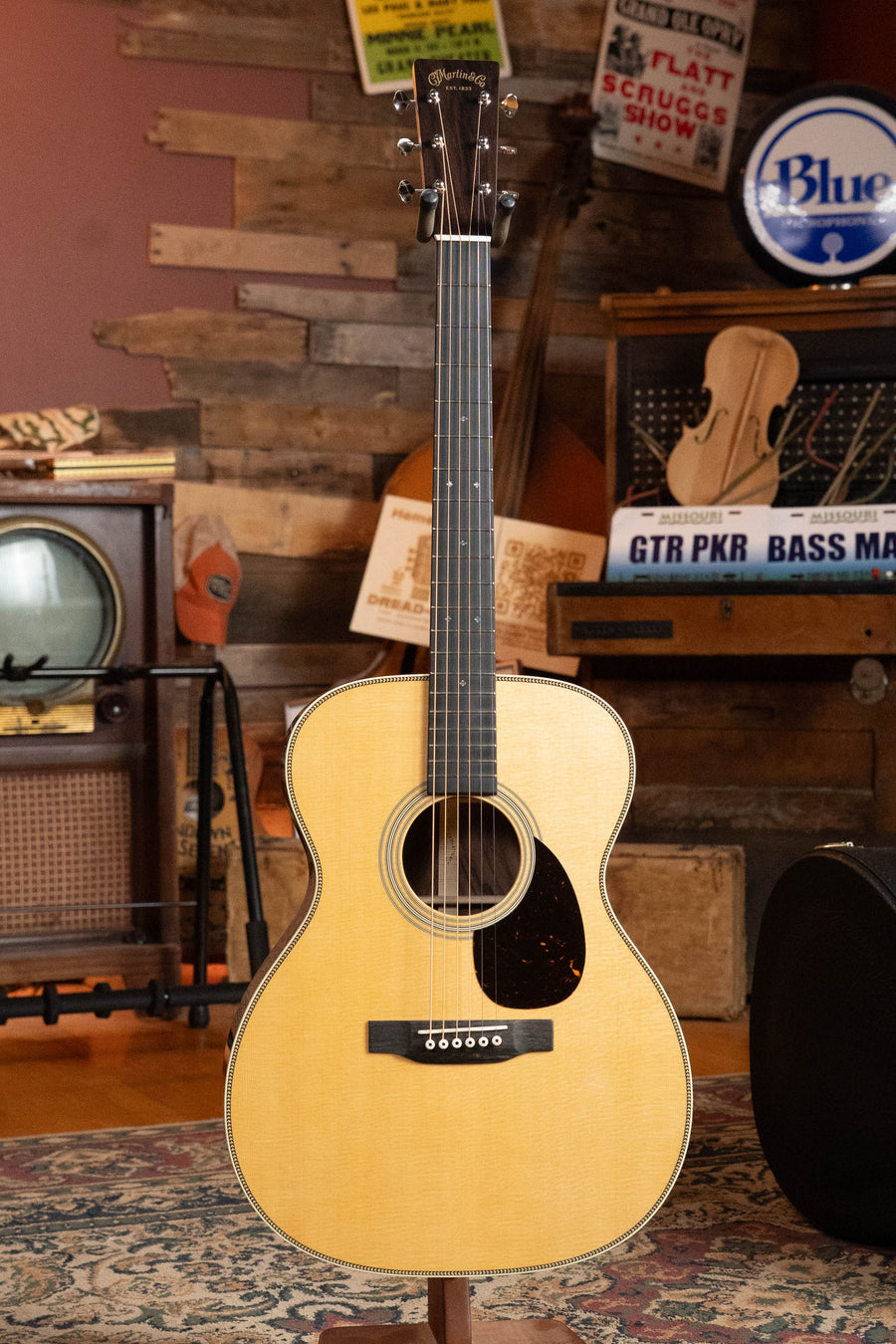 A Martin & Co. Custom Shop OOO-28 Style "Wild Grain" East Indian Rosewood Auditorium Acoustic Guitar #6894 with a natural wood finish stands in a room adorned with vintage instruments, amps, signs, and rustic wood panels.