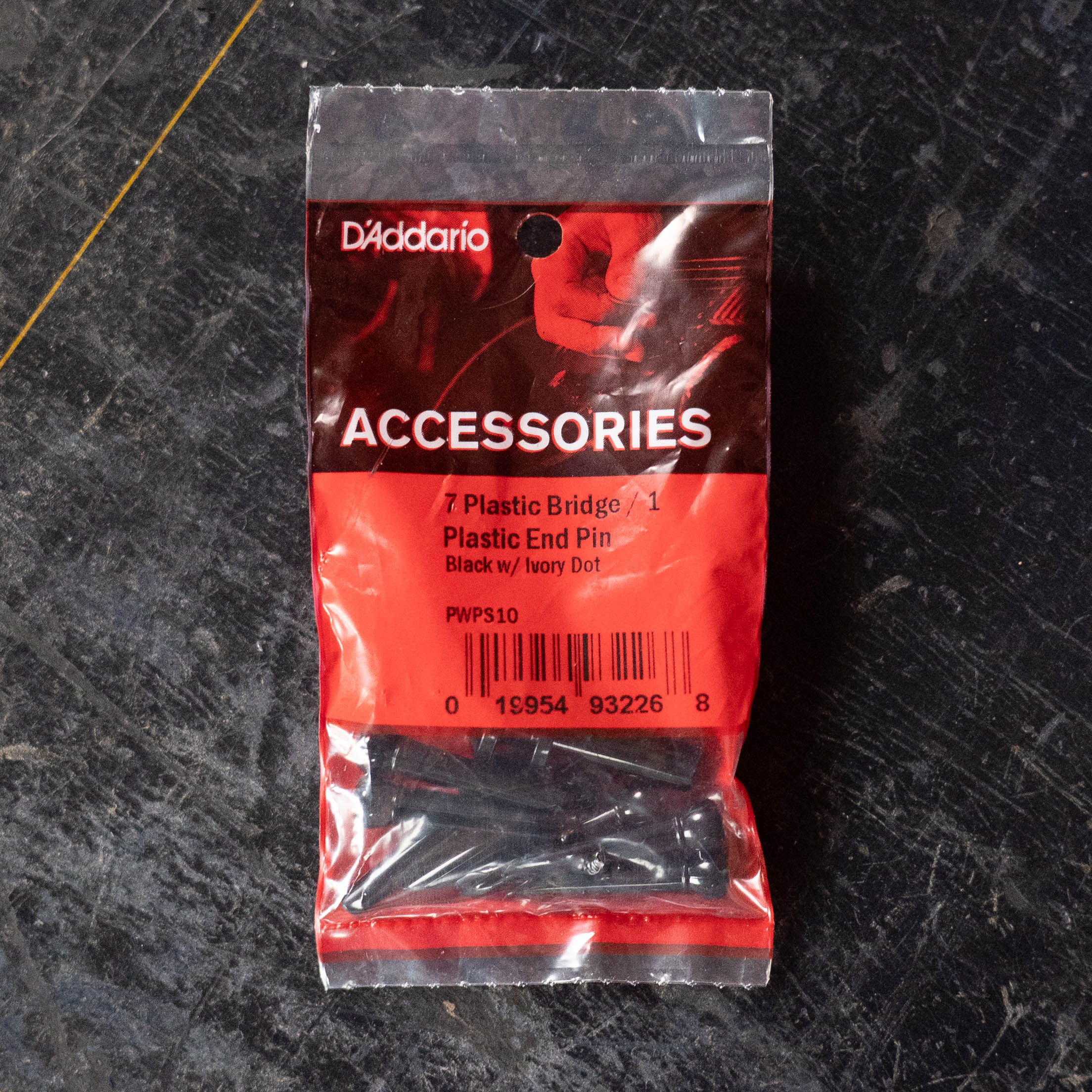 A sealed pack of D'Addario Injected Molded Bridge Pins w/End Pin Set, Ebony with Ivory Dot (PWPS10), sits on a dark textured surface. The acoustic guitar bridge pins are visible through the packaging.