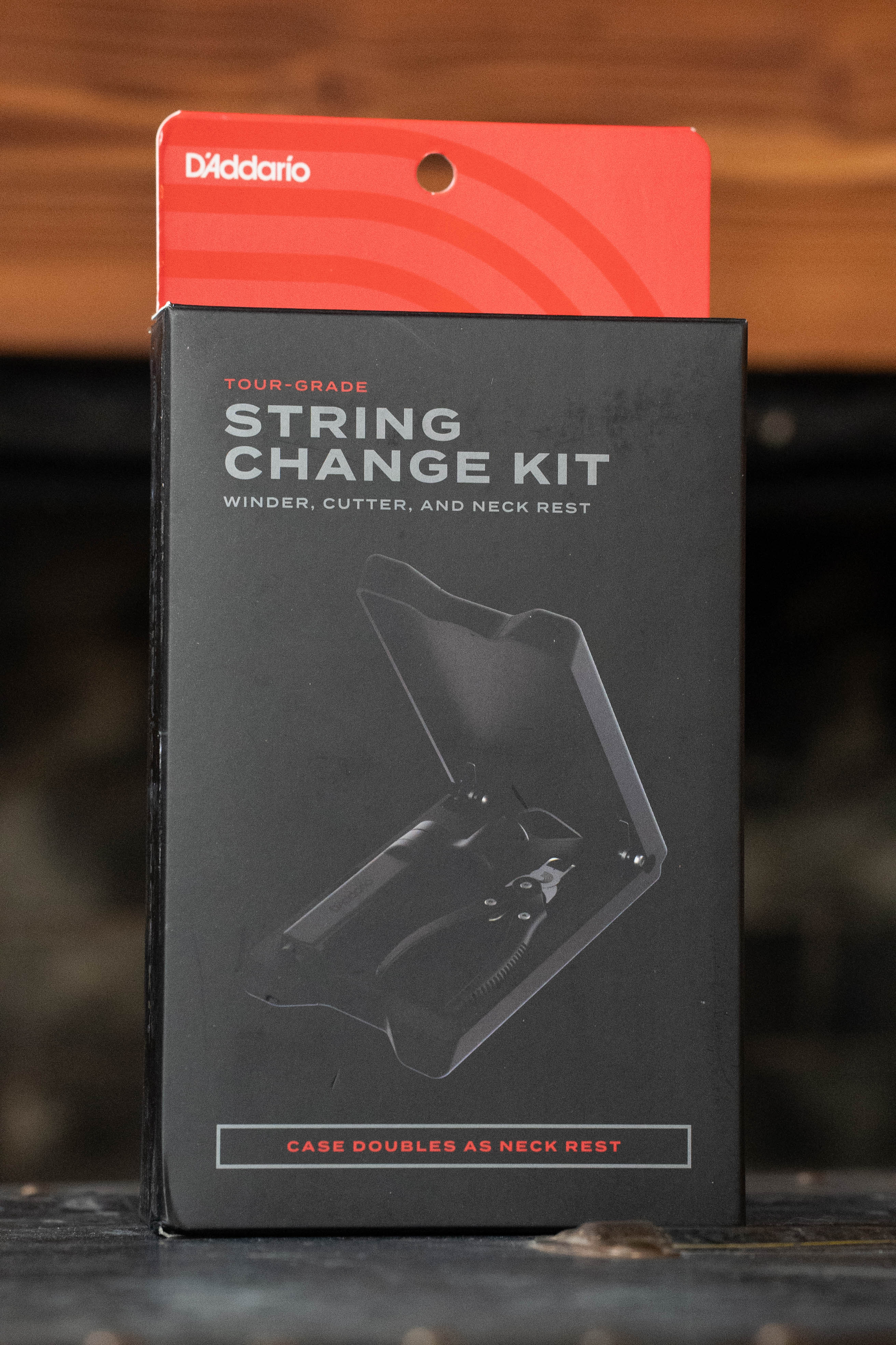 A box of the D'Addario Tour Grade String Changing Kit, displaying the kit image and text noting its use as a winder, string cutter, bridge pin puller, and neck rest. The black packaging features red accents. Brand: D'Addario.