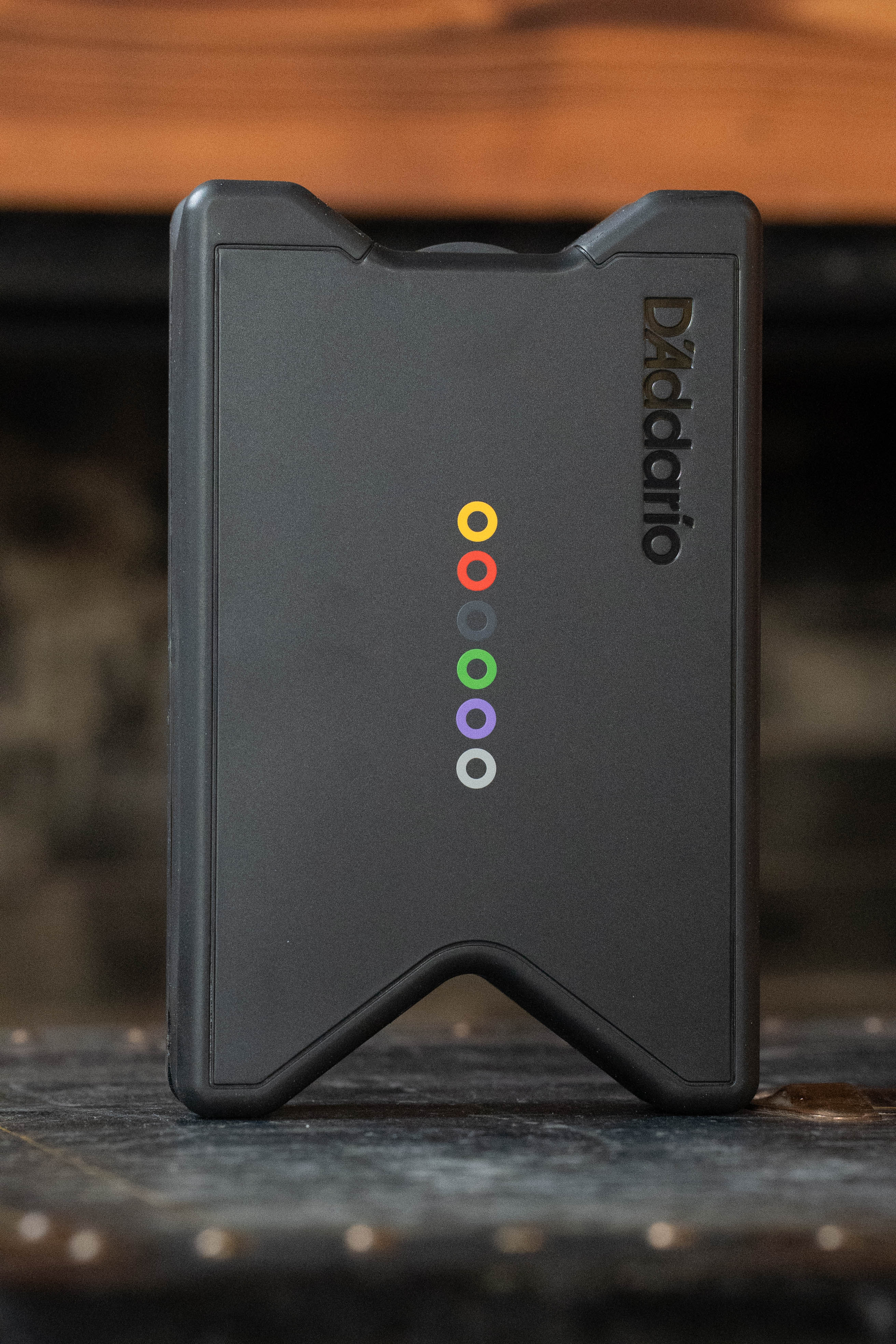 A black D’Addario Tour Grade String Changing Kit box stands upright on a wooden surface, featuring six colored circles vertically in the center and the D’Addario brand, with a blurred background.