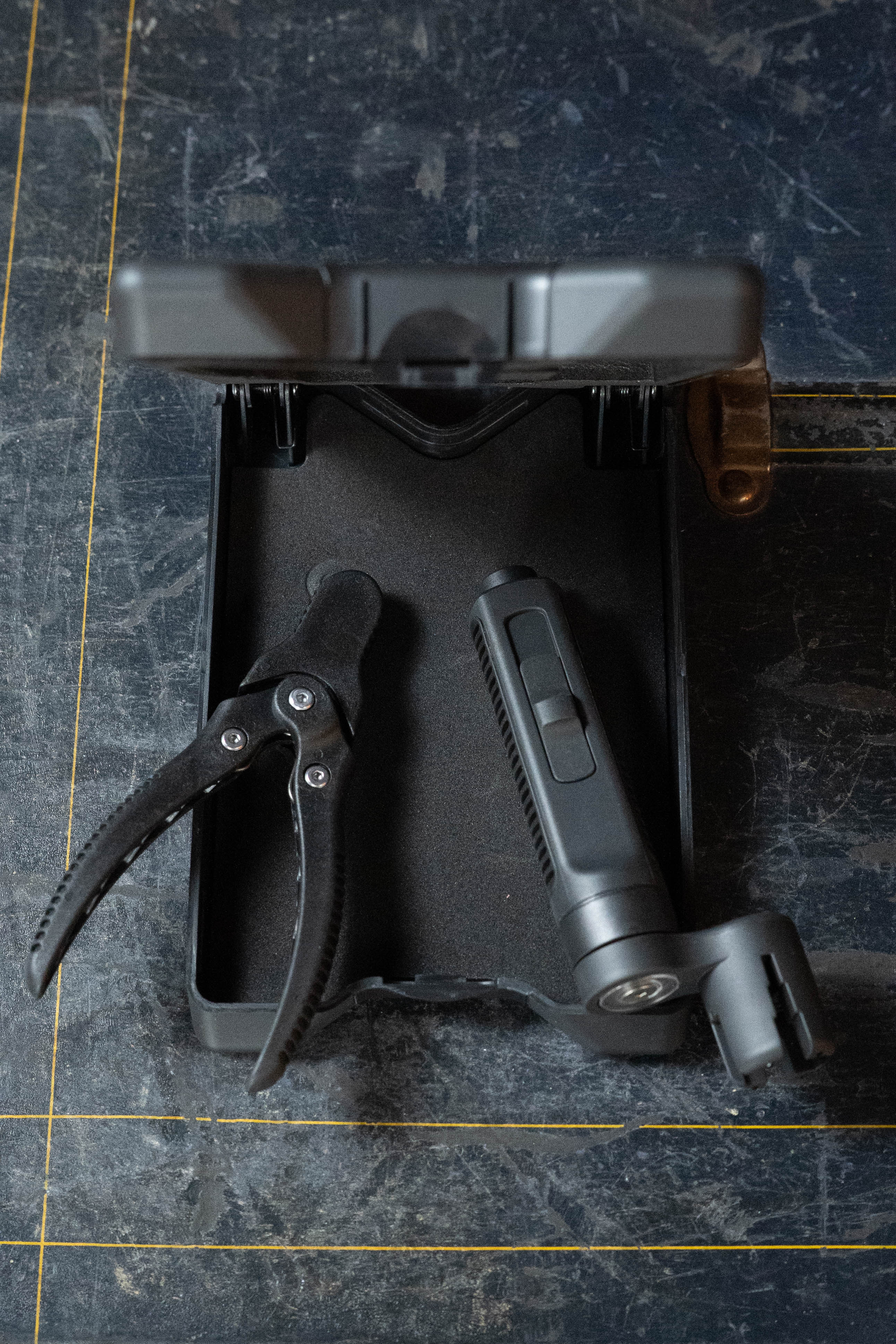 The D'Addario Tour Grade String Changing Kit by D'Addario is displayed in an open black case on a dark surface, showing two black spring clamps, a string cutter with a ridged handle, and yellow measuring lines on the surface.