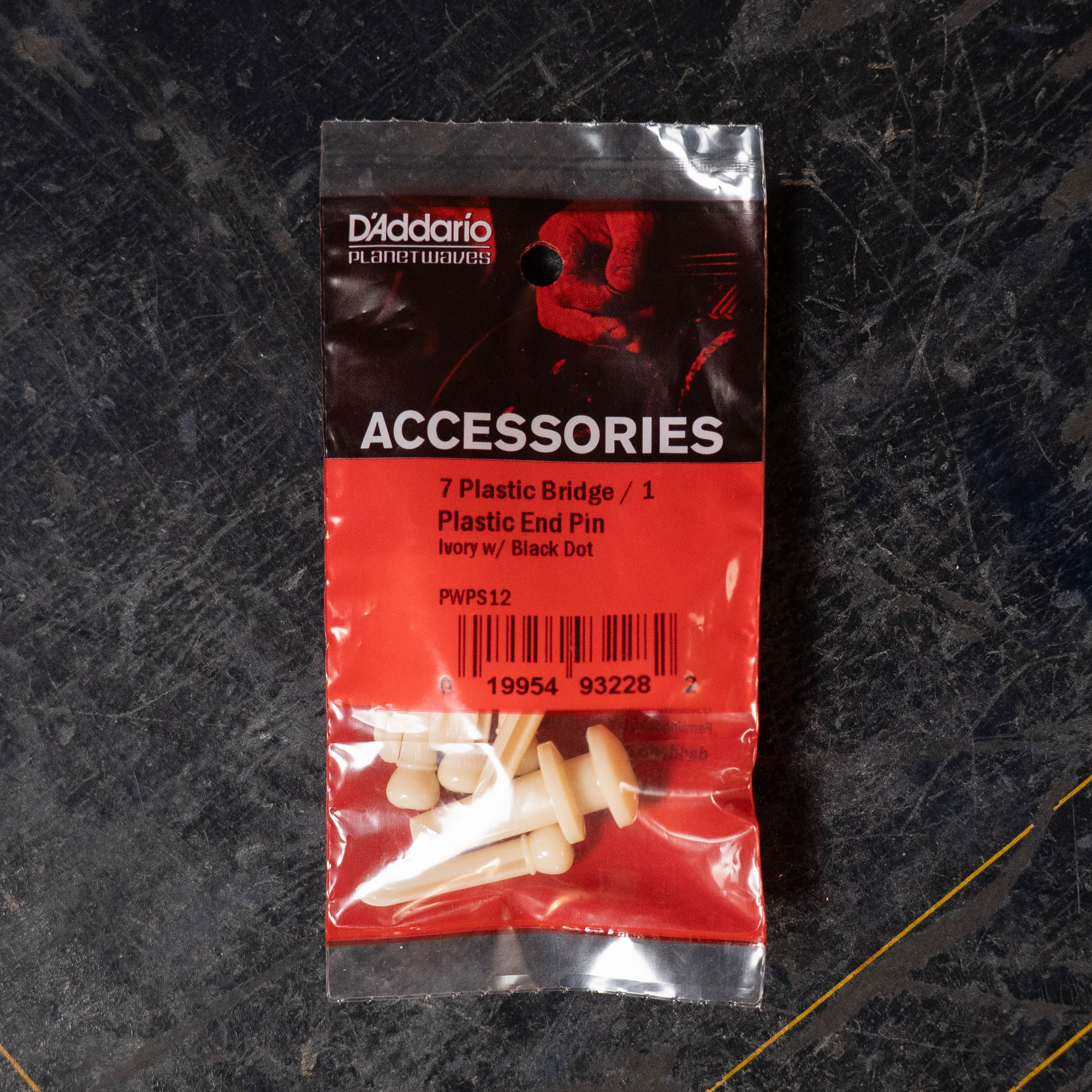 A D'Addario Injected Molded Bridge Pins with End Pin, Set of 7 - Ivory, includes seven ivory plastic acoustic guitar bridge pins with black dots from D'Addario, shown on a dark textured surface.