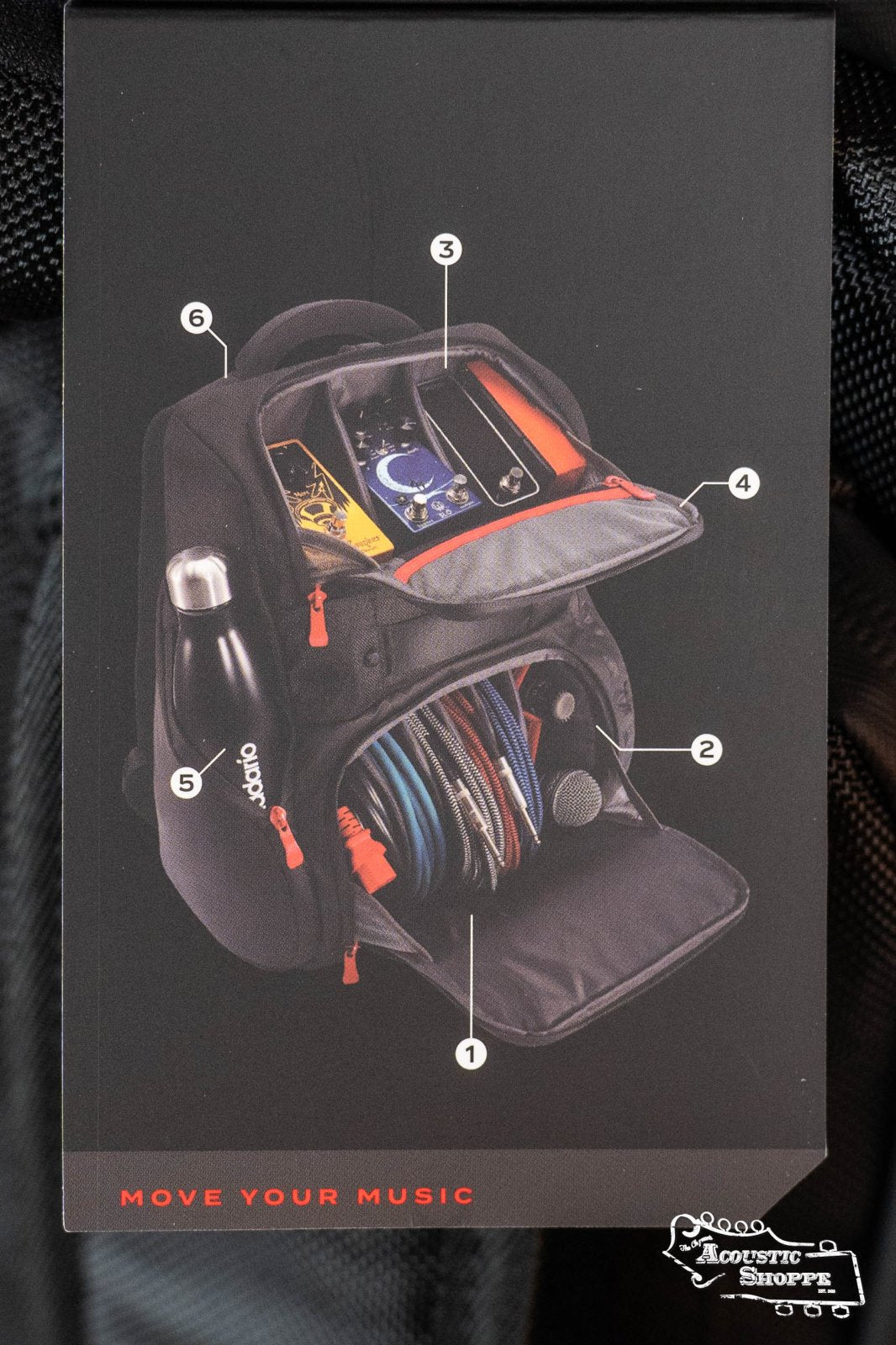 The DAddario Backline Gear Transport Pack Solo, a black musician backpack with labeled compartments for water bottle, cables, microphone, phone, and music gear, is open to show organized audio storage. Move Your Music appears below. Brand: DAddario.