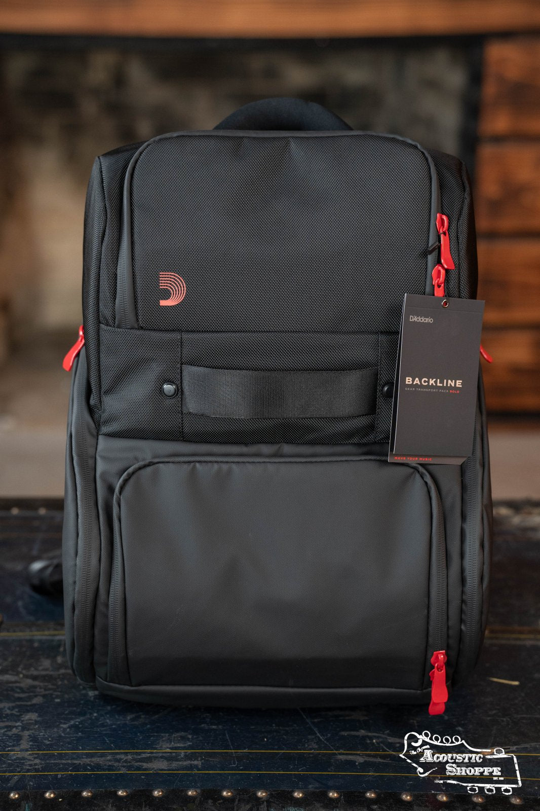 The DAddario Backline Gear Transport Pack Solo is a black musicians backpack with red zipper pulls and a front pocket, shown upright on wood. The BACKLINE tag is visible, with a blurred fireplace and furniture in the background.