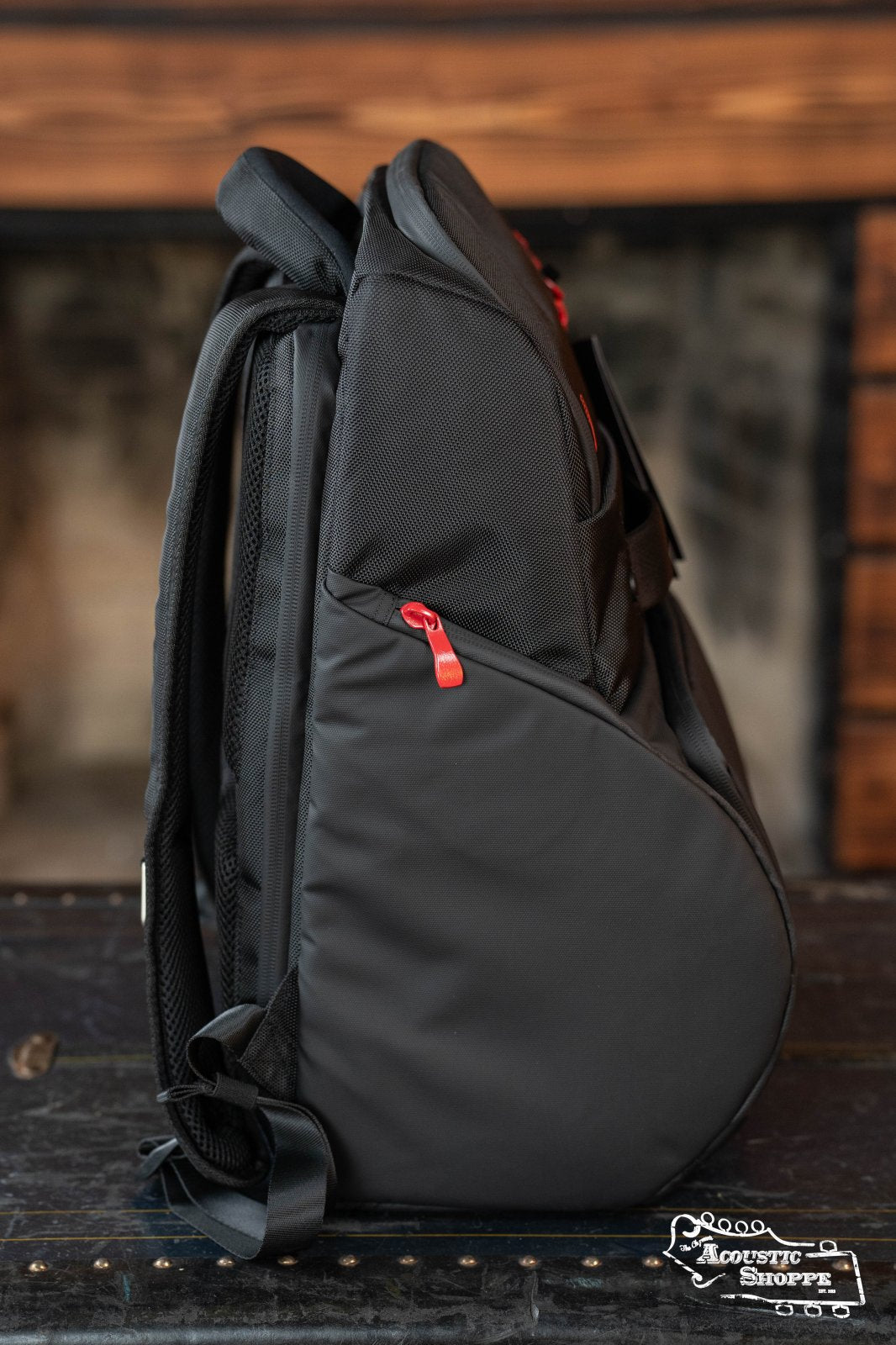 The DAddario Backline Gear Transport Pack Solo stands upright on a dark wooden surface, showcasing black fabric, red zipper pulls, padded straps, and a slanted side pocket. A blurred fireplace and mantel appear in the background.