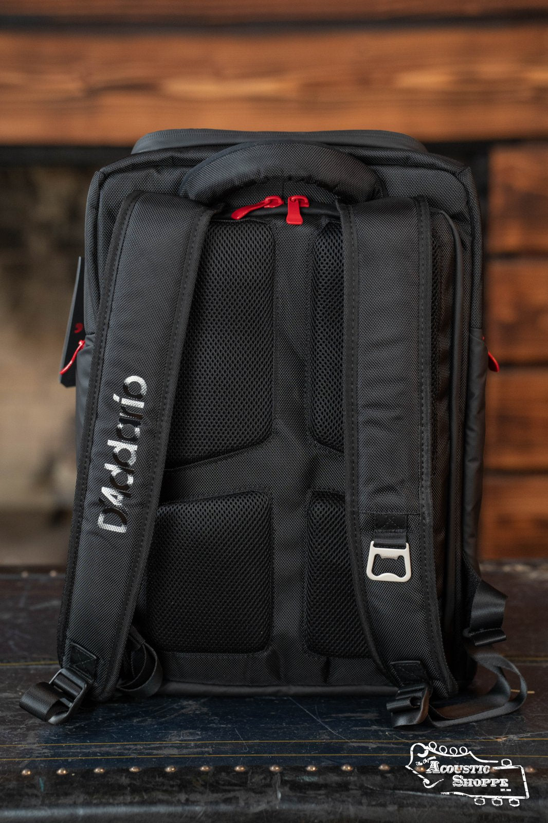 A DAddario Backline Gear Transport Pack Solo backpack with padded straps and red zipper pulls sits upright on a dark surface against a blurred wooden background. The Acoustic Shoppe logo appears in the bottom right corner.