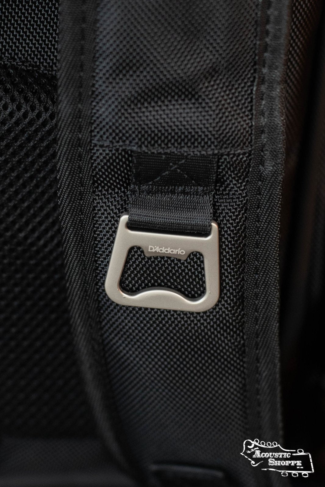 Close-up of the DAddario Backline Gear Transport Pack Solo shows a metal DAddario-branded bottle opener on the textured strap, with part of The Acoustic Shoppe logo, showcasing this gig bags professional and thoughtful design.