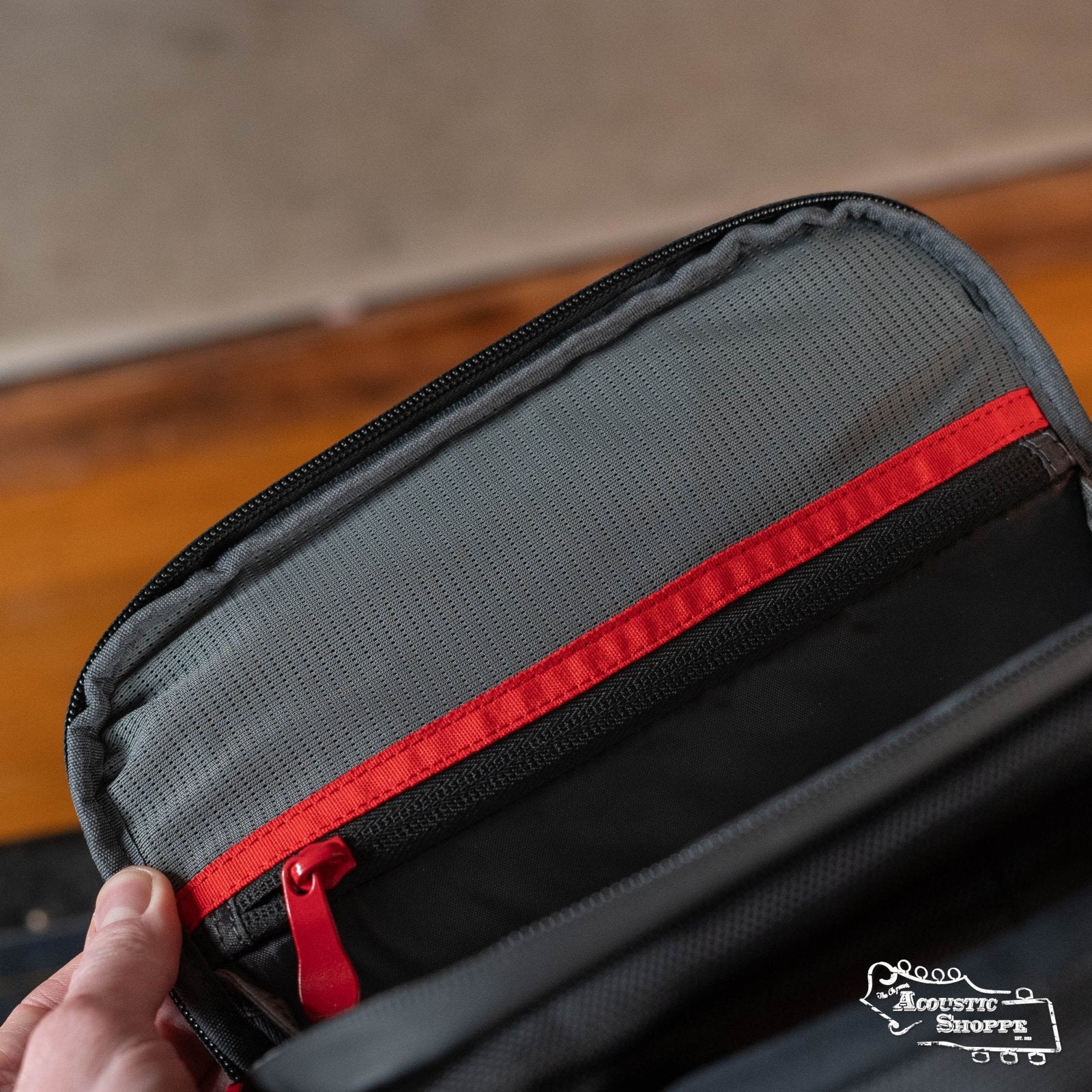 A close-up of a DAddario Backline Gear Transport Pack Solo backpack pocket in gray and black with a red zipper. A hand holds the open pocket, and part of a wooden floor is visible in the background.