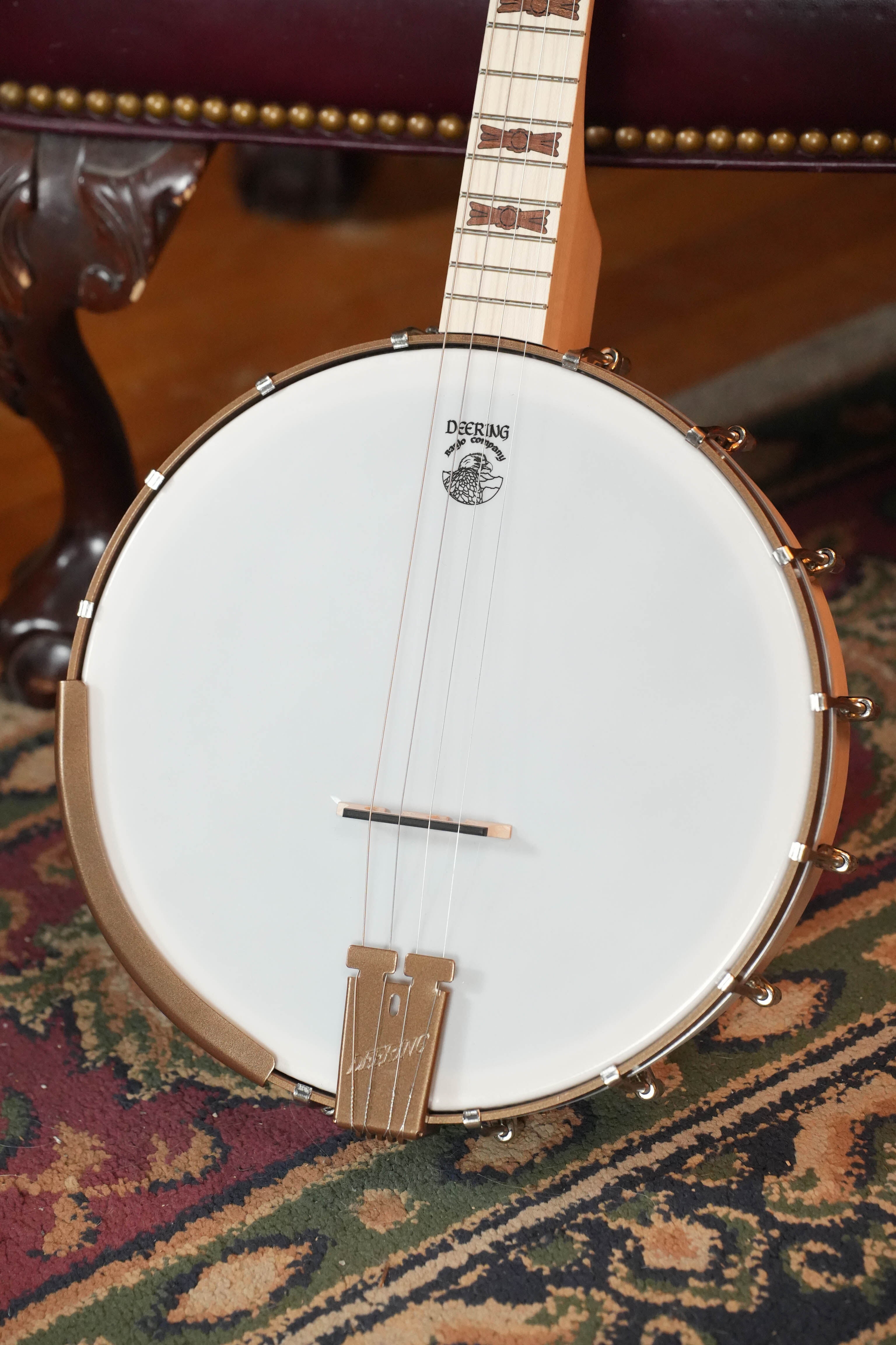 A close-up of the Deering Goodtime 19-Fret Tenor Open Back Banjo #92620 by Deering Banjos, lightweight with a maple neck, drumhead, bridge, tailpiece, and inlaid fretboard, rests on a patterned rug beside a wooden chair leg.