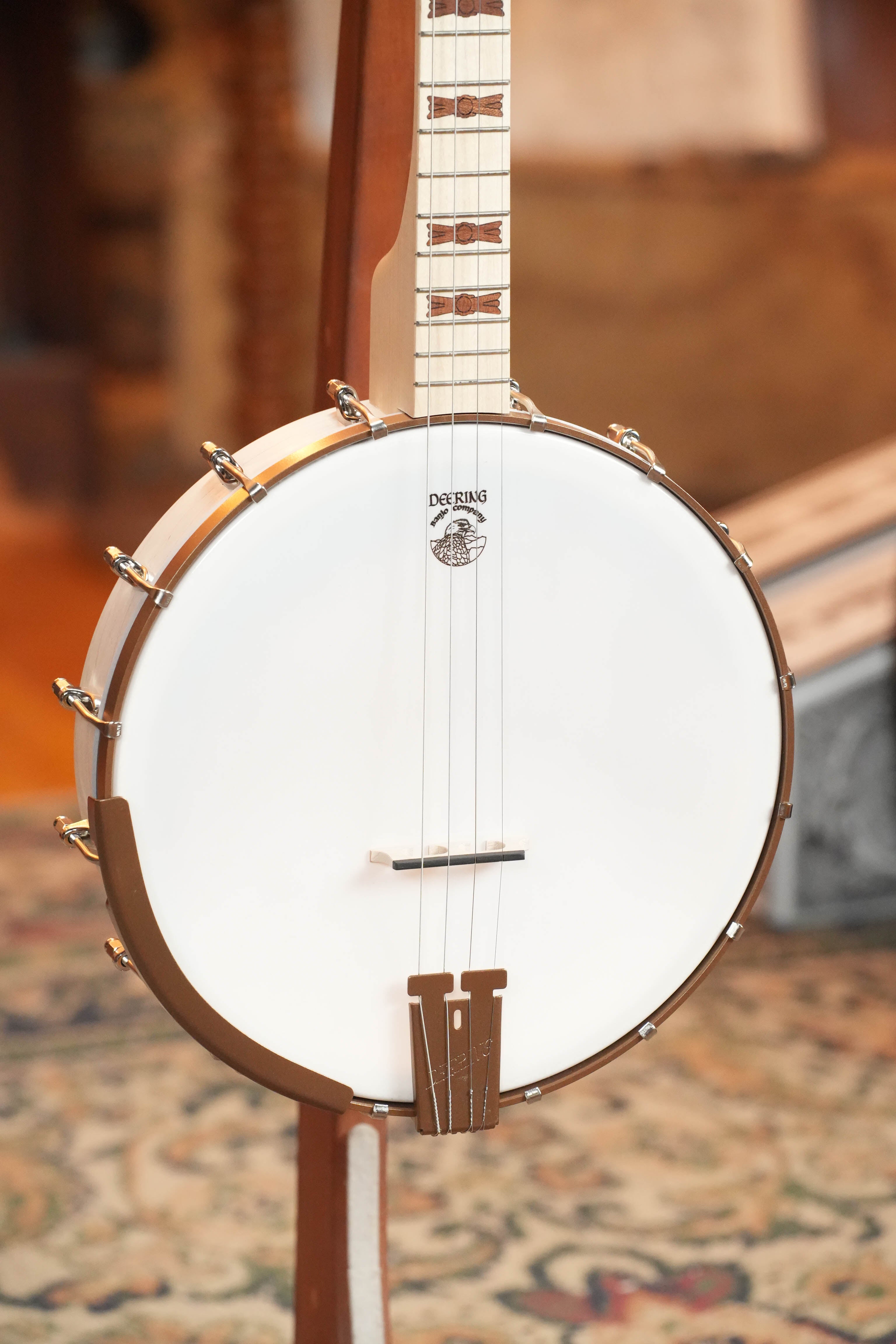 A close-up of a Deering Goodtime 19-Fret Tenor Open Back Banjo #92620 by Deering Banjos, featuring a maple neck, white drumhead, metallic hardware, and light wood finish, resting on a stand atop a patterned rug.
