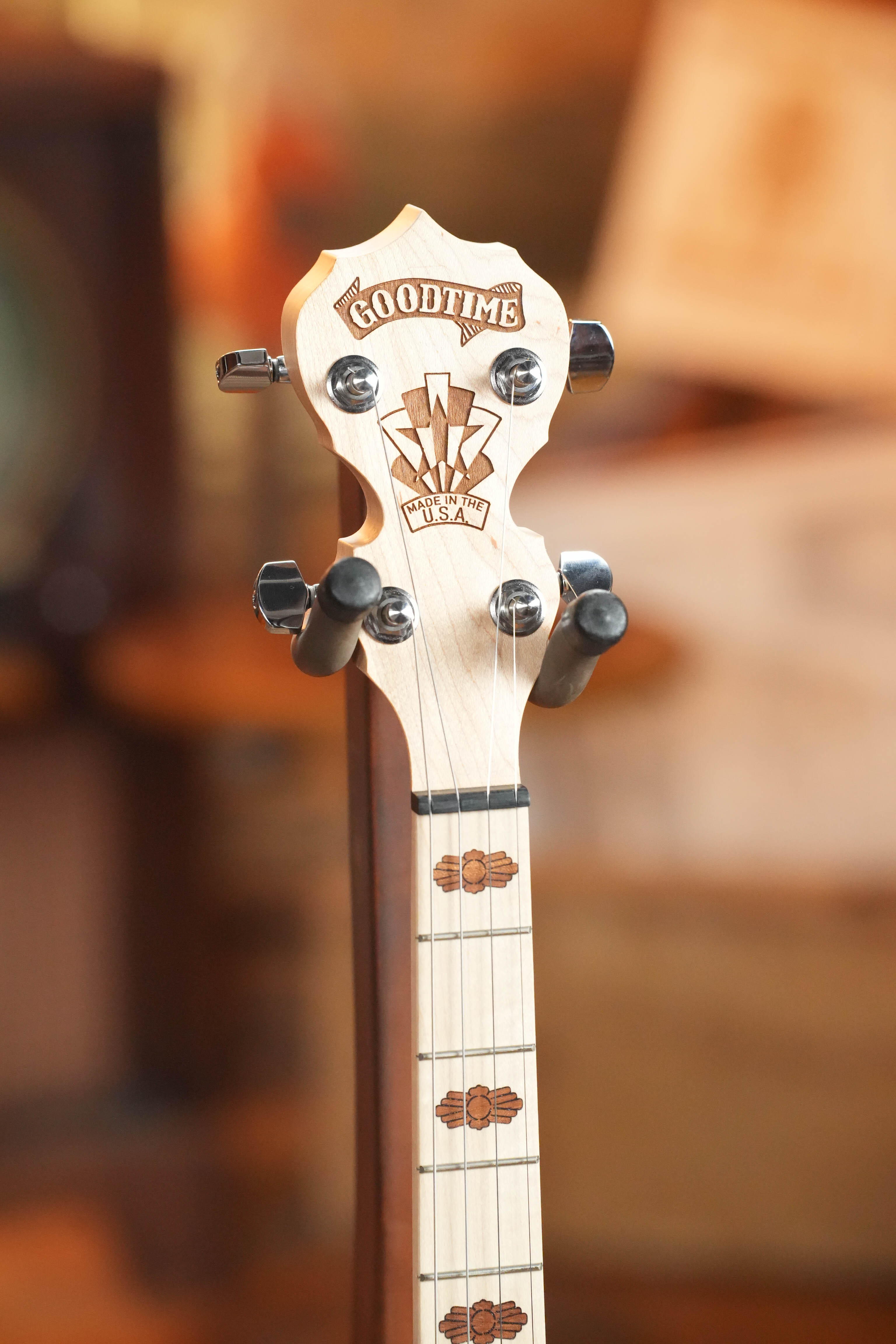 Close-up of the Deering Goodtime 19-Fret Tenor Open Back Banjo #92620 headstock by Deering Banjos, showing tuning pegs, engraved logo, and decorative inlays on maple wood against a blurred background.