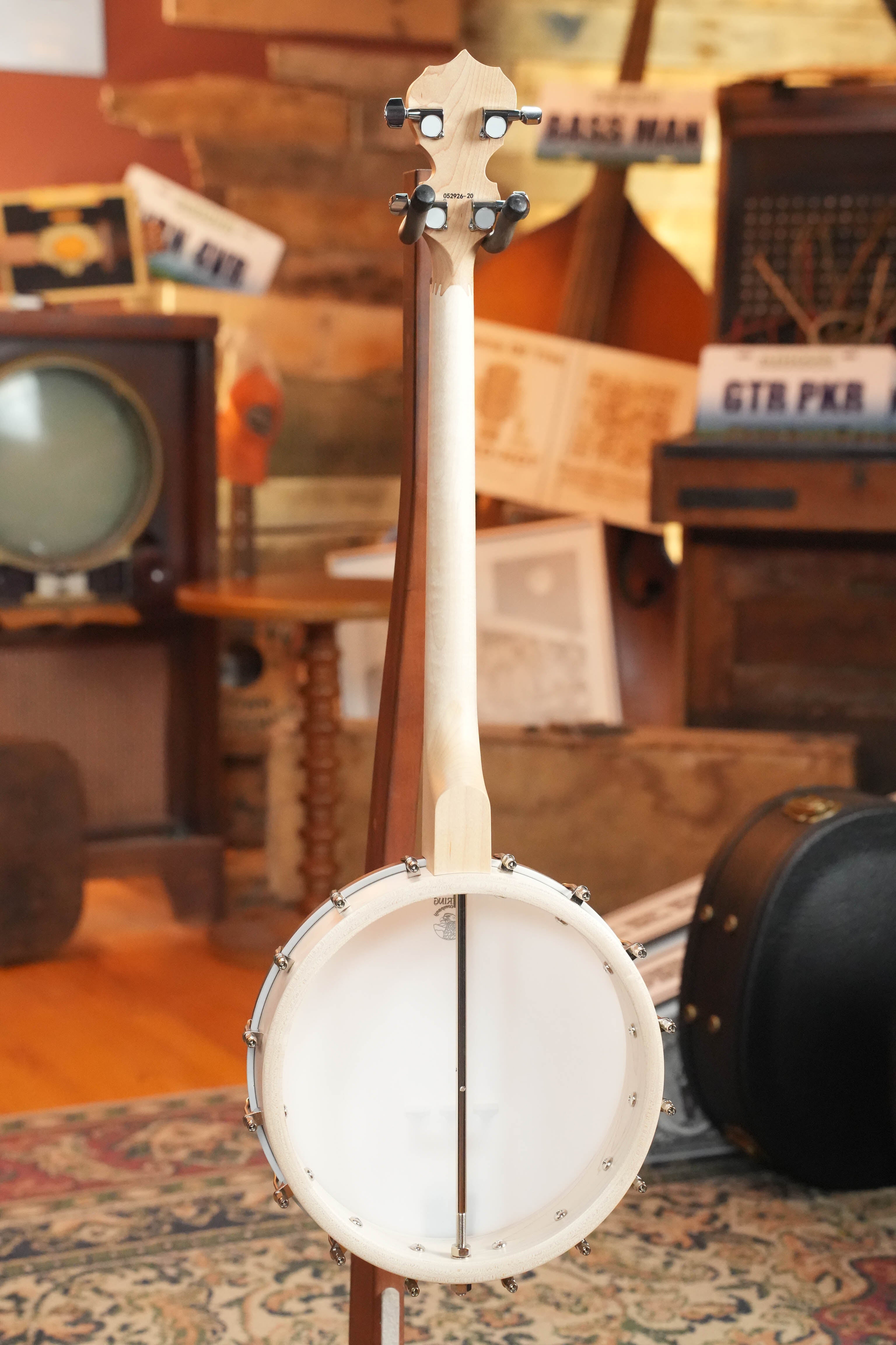 The Deering Goodtime 19-Fret Tenor Open Back Banjo #92620 by Deering Banjos, a lightweight white banjo with a maple neck, is displayed on a stand in a cozy room filled with vintage instruments and music-themed decor.