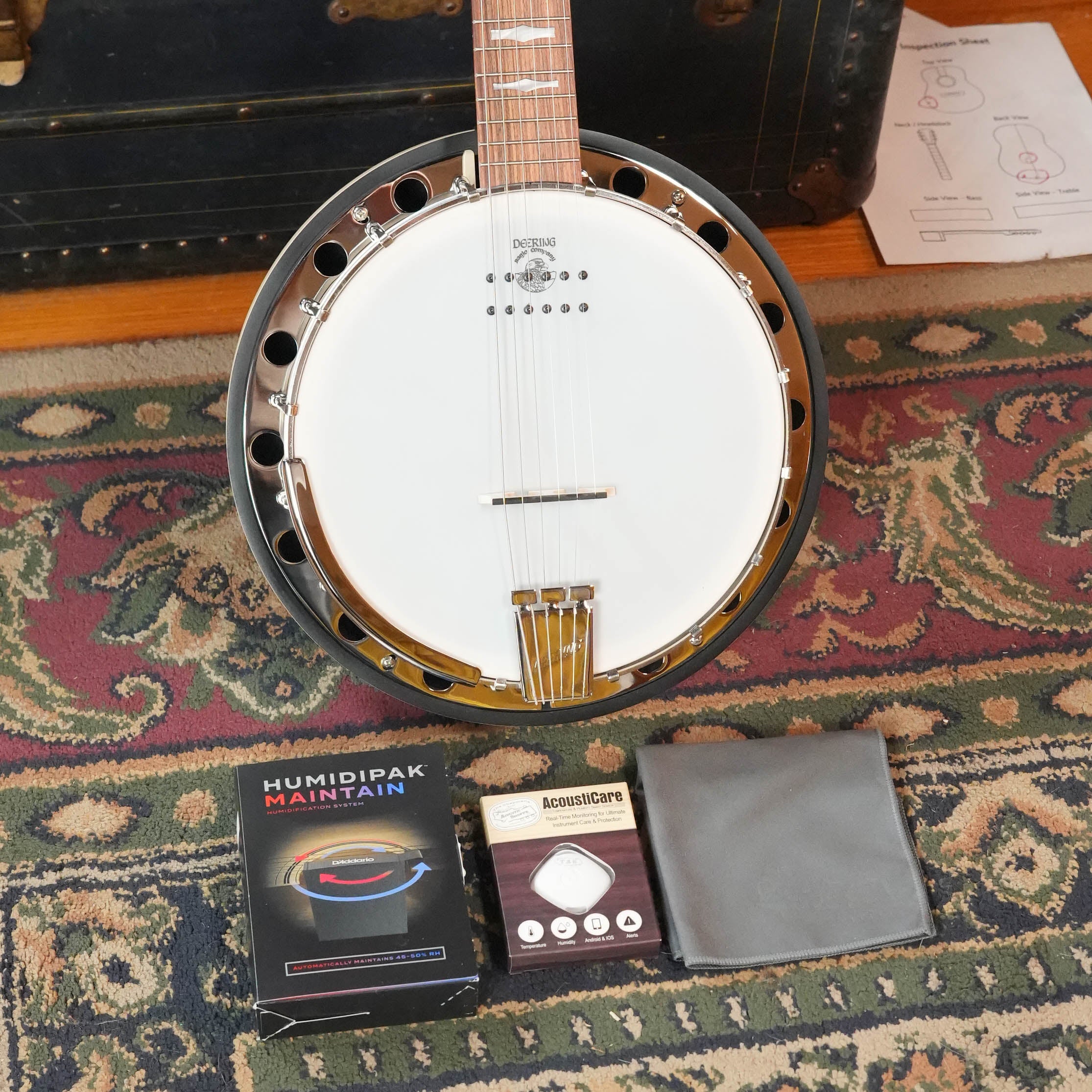 A Deering Banjos Goodtime 6 R Steel String Banjo with Kavanjo Pickup (#2620) rests on a patterned rug, with a Humidipak Maintain pack, hygrometer, and gray cloth in front. An old trunk and papers appear in the background.