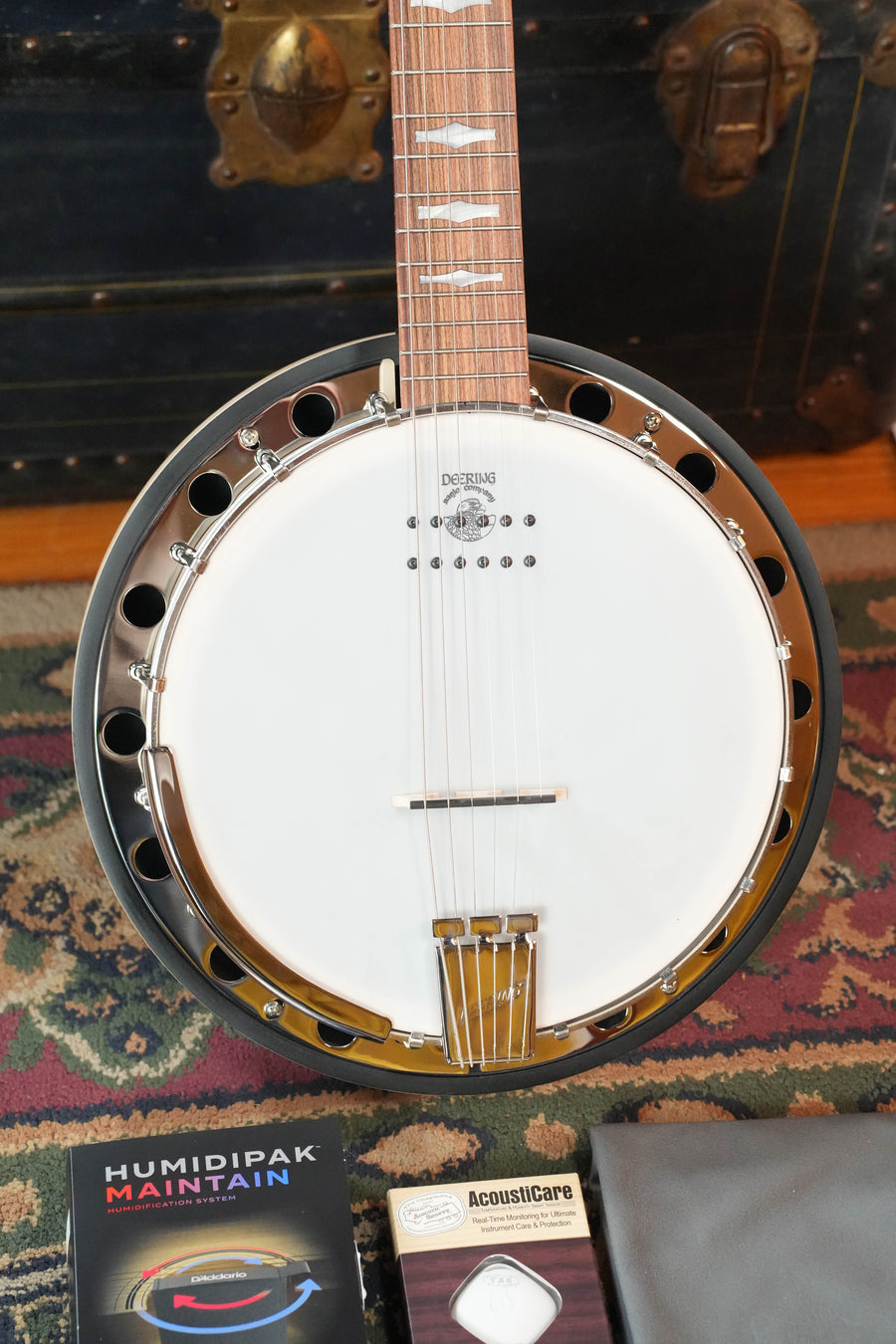 A Deering Banjos Goodtime 6 R Steel String Banjo with Kavanjo Pickup rests on a patterned rug. Nearby are a Humidipak Maintain package, an AcoustiCare item, and a gray cloth. A vintage trunk sits in the background.