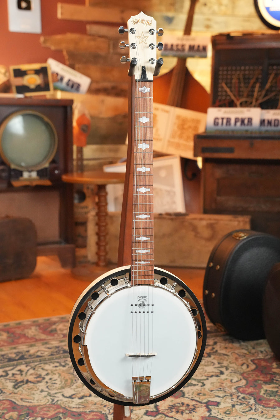 The Deering Goodtime 6 R Steel String Banjo W/Kavanjo Pickup #2620 by Deering Banjos stands upright in a room filled with vintage musical and television equipment.