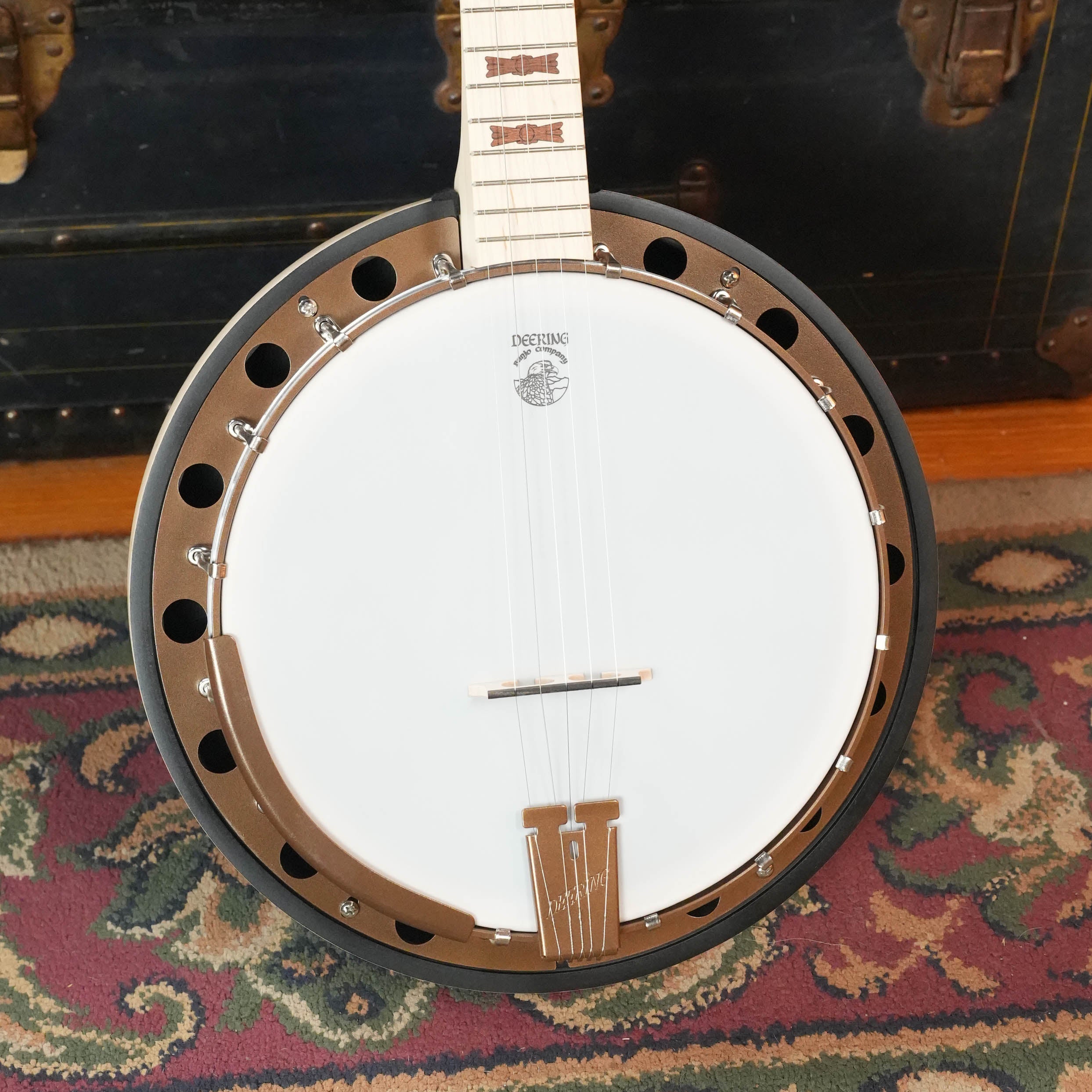A close-up of the Deering Goodtime Two 5 String Resonator Banjo #2615 by Deering Banjos resting on a patterned rug highlights its white drum head, strings, and detailed metal rim, with a dark vintage trunk in the background.