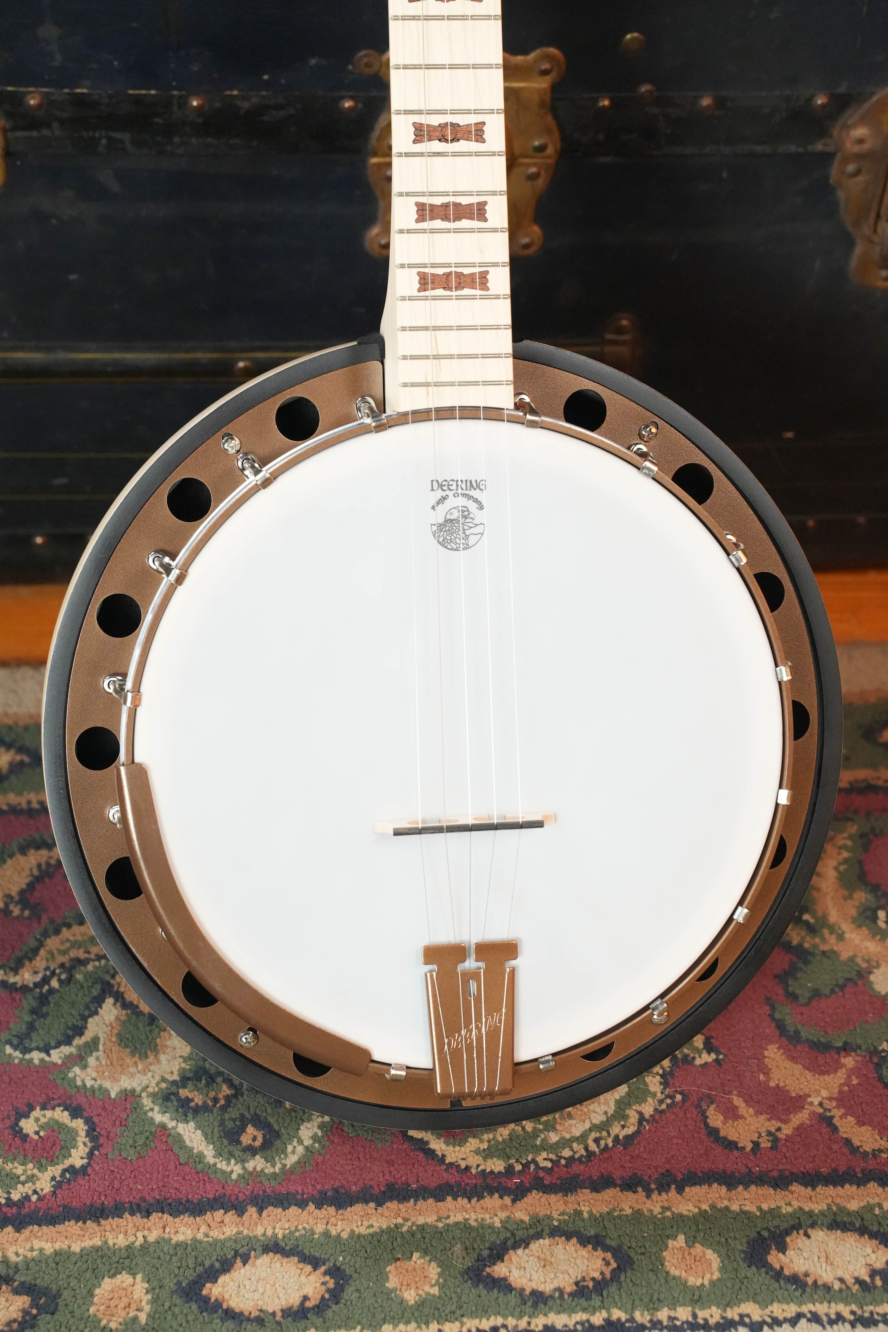 A close-up of the Deering Banjos Goodtime Two 5 String Resonator Banjo #2615 upright on a patterned rug, highlighting its white drumhead, metallic rim with circular cutouts, and neck with intricate inlays.