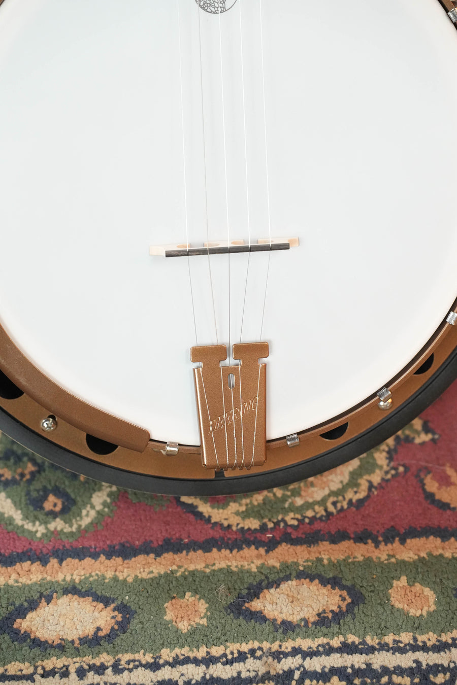 Close-up of a Deering Goodtime Two 5 String Resonator Banjo #2615 by Deering Banjos, showing its body, tailpiece, strings, bridge, and part of the banjo head resting on a colorful patterned rug.