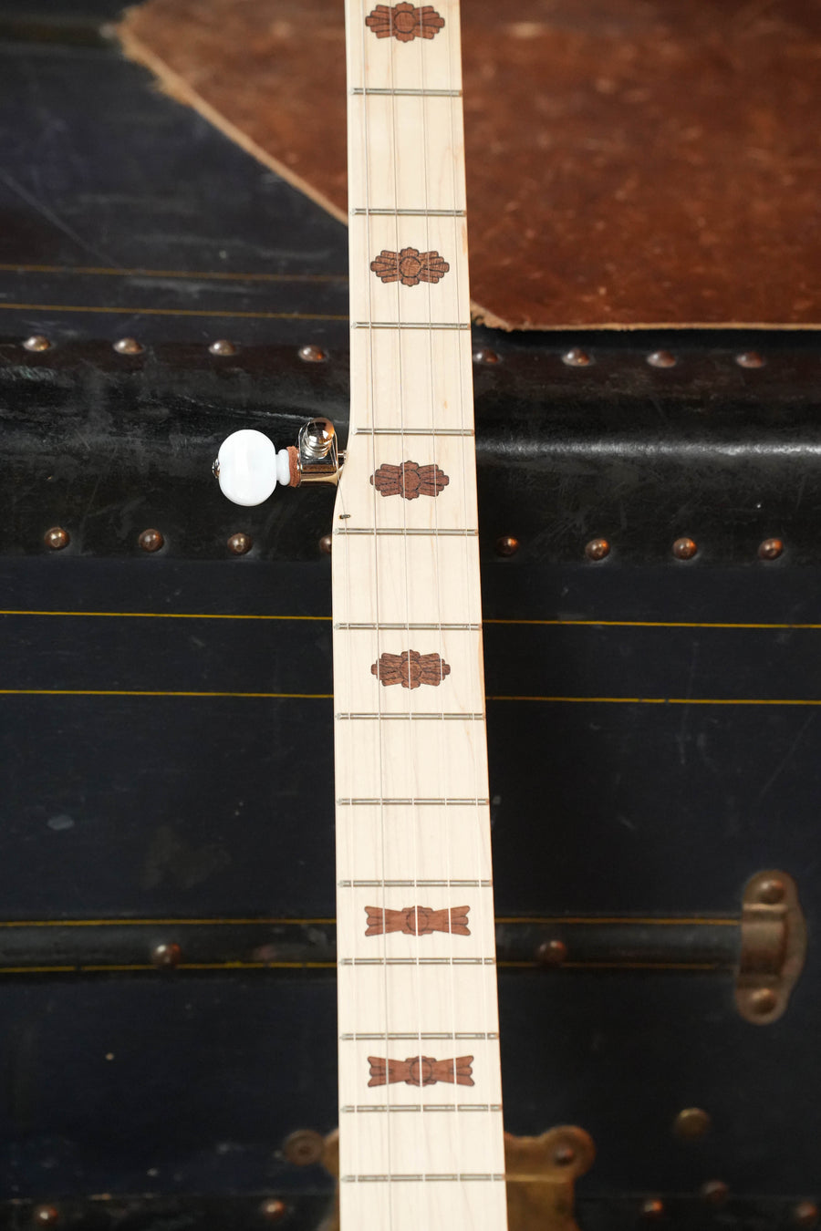 A close-up of the Deering Goodtime Two 5 String Resonator Banjo #2615 by Deering Banjos, showing decorative inlays on the neck, set against an old black trunk with metal rivets and leather details.