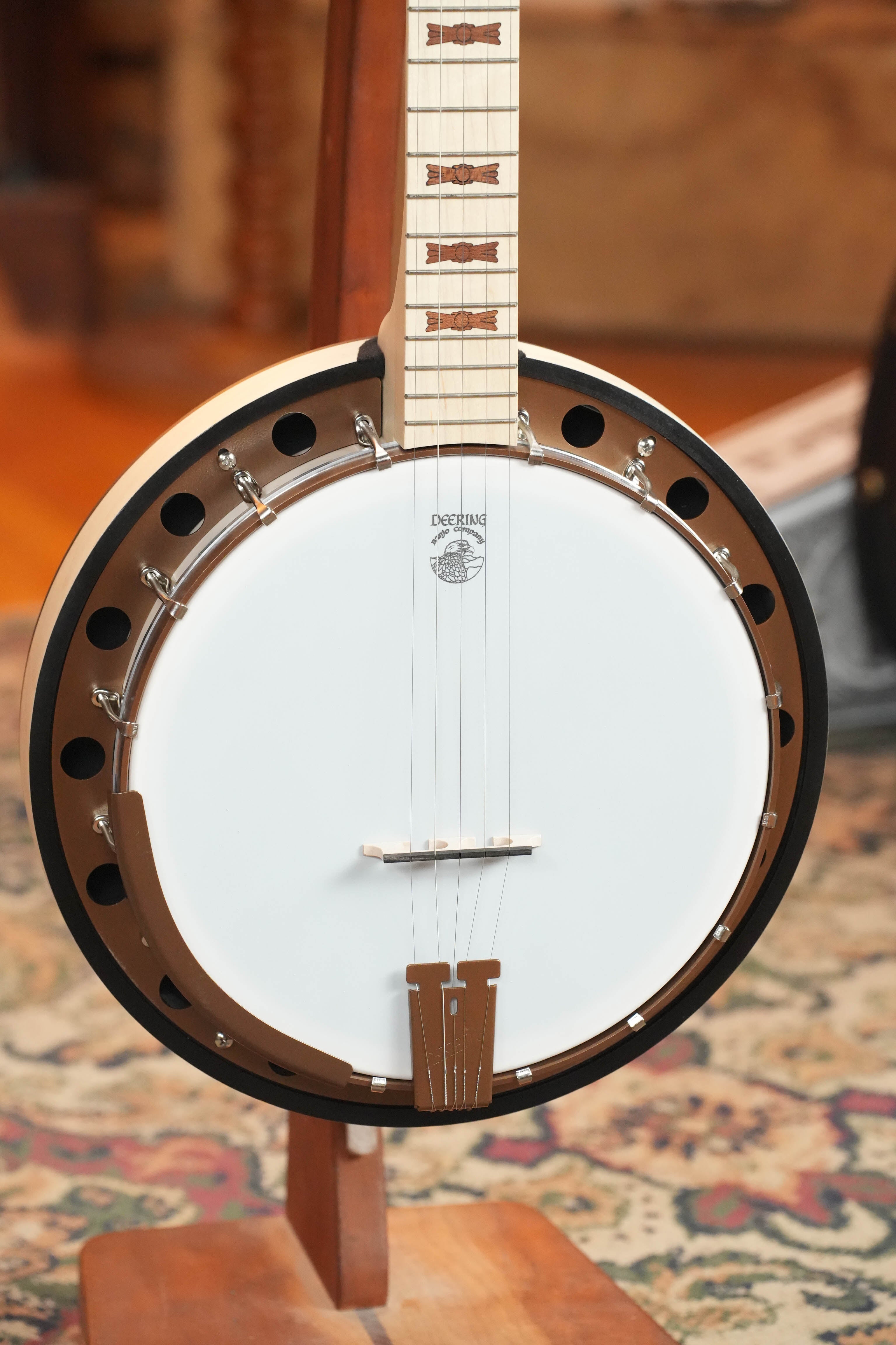 A close-up of the Deering Banjos Goodtime Two 5 String Resonator Banjo #2615 on a stand, highlighting its white drumhead, metal rim with holes, strings, neck with inlays, plus a patterned rug and wooden floor in the background.
