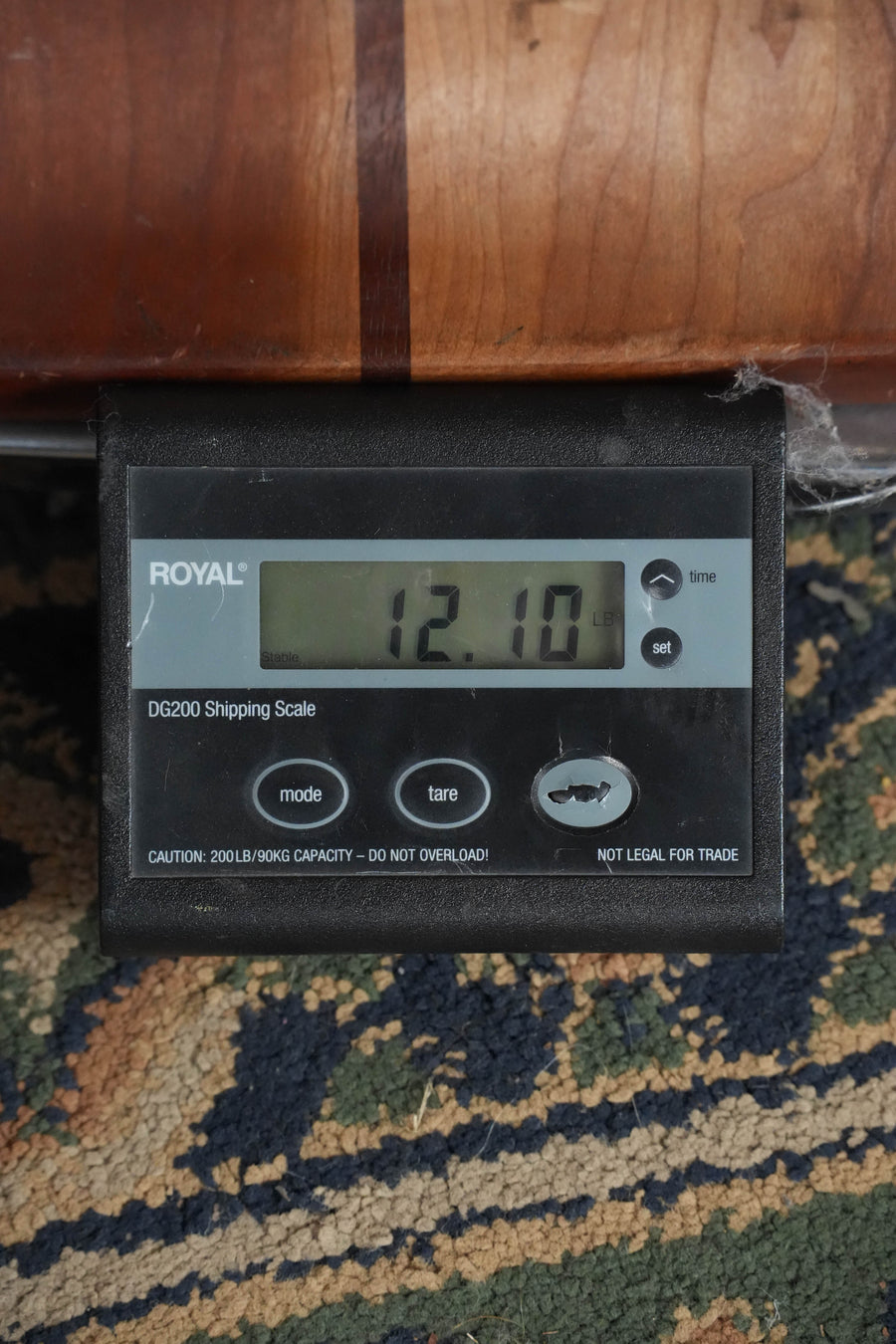 A digital shipping scale labeled ROYAL DG200 reads 12.10 lbs on a patterned carpet, with the Deering Julia Belle 5 String Resonator Banjo w/Spikes Installed (#0067) by Deering Banjos partially visible above it.