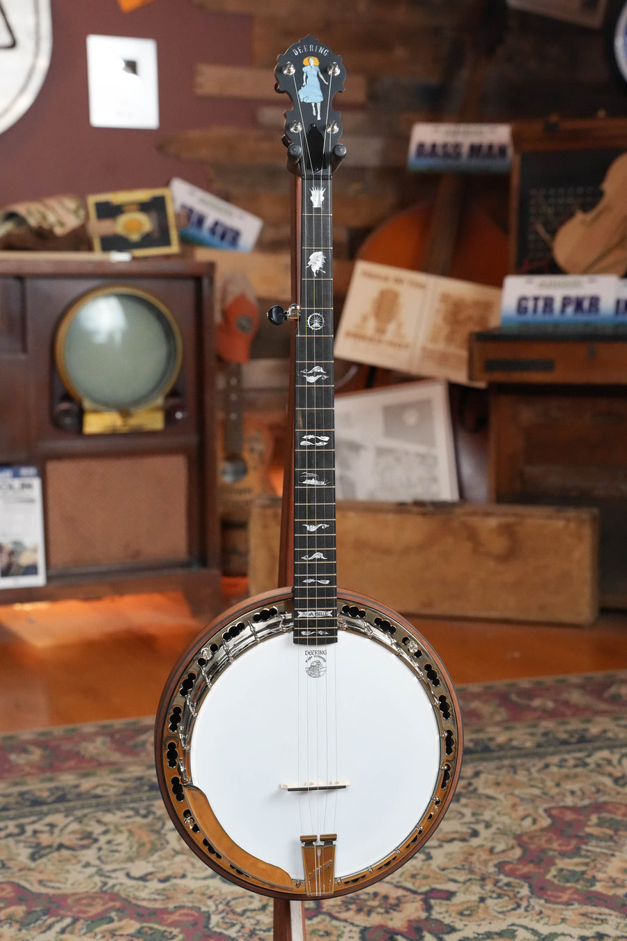 A Deering Banjos Julia Belle 5 String Resonator Banjo w/Spikes Installed (#0067) stands on a patterned rug in a vintage room featuring wood paneling, a retro TV, musical instruments, and nostalgic photos. For sale.