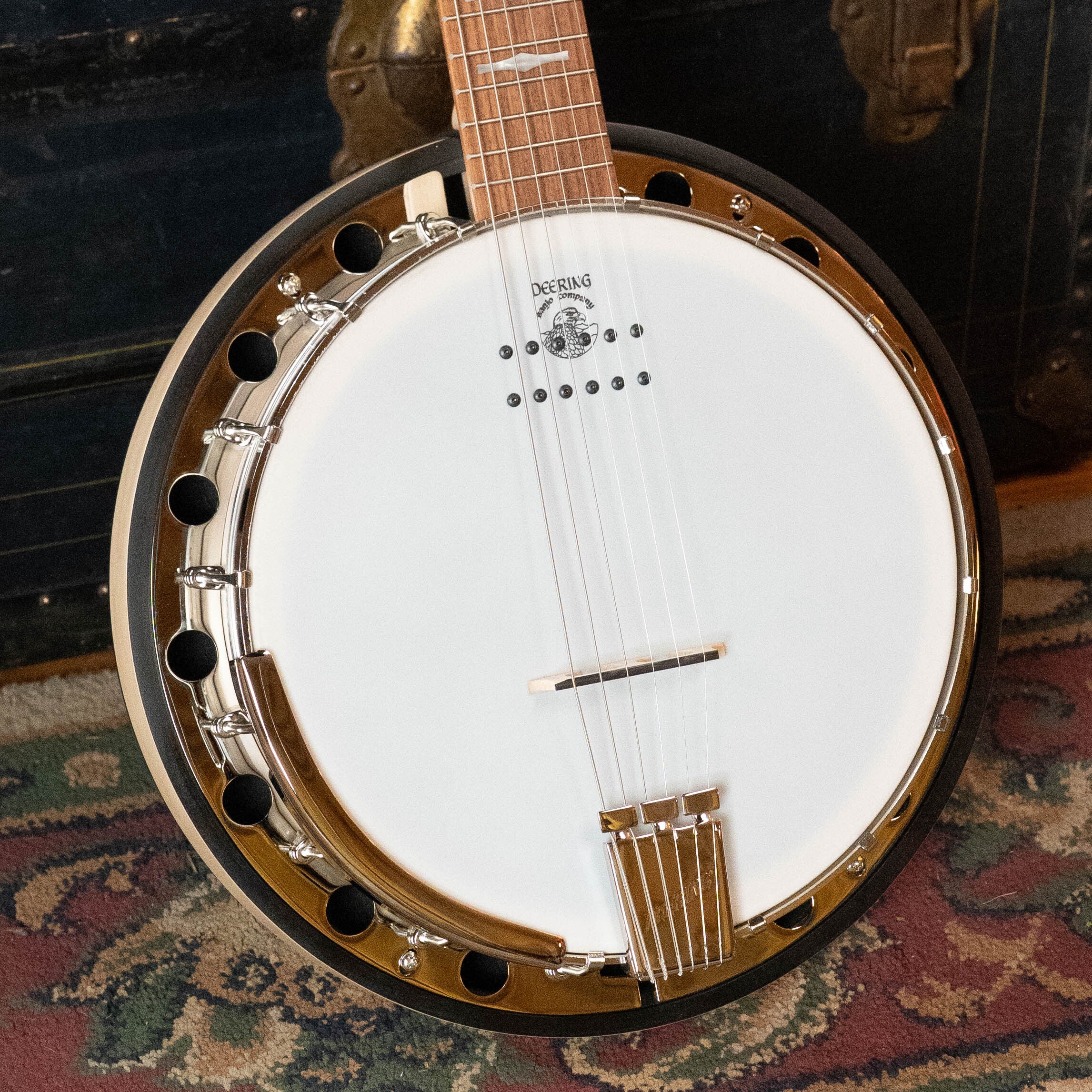 Close-up of the Deering Goodtime Six-R 6-String Resonator Banjo w/Kavanjo Pickup by Deering Banjos on a patterned rug, displaying its drumhead, metallic hardware, wooden neck, and strings with a dark brass-trimmed trunk in the background.